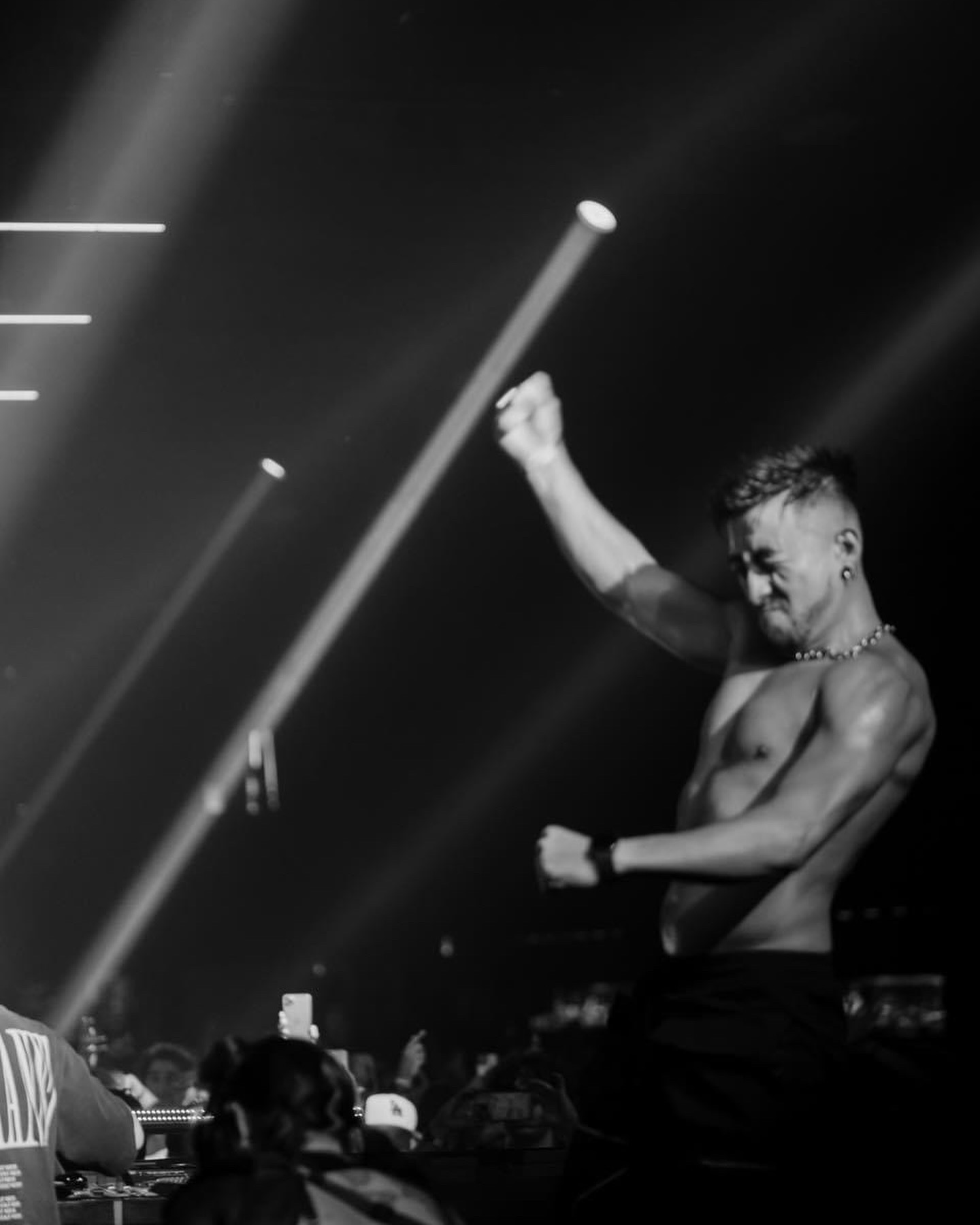 A shirtless male performer on stage with one arm raised and the other bent, surrounded by stage lights and audience.