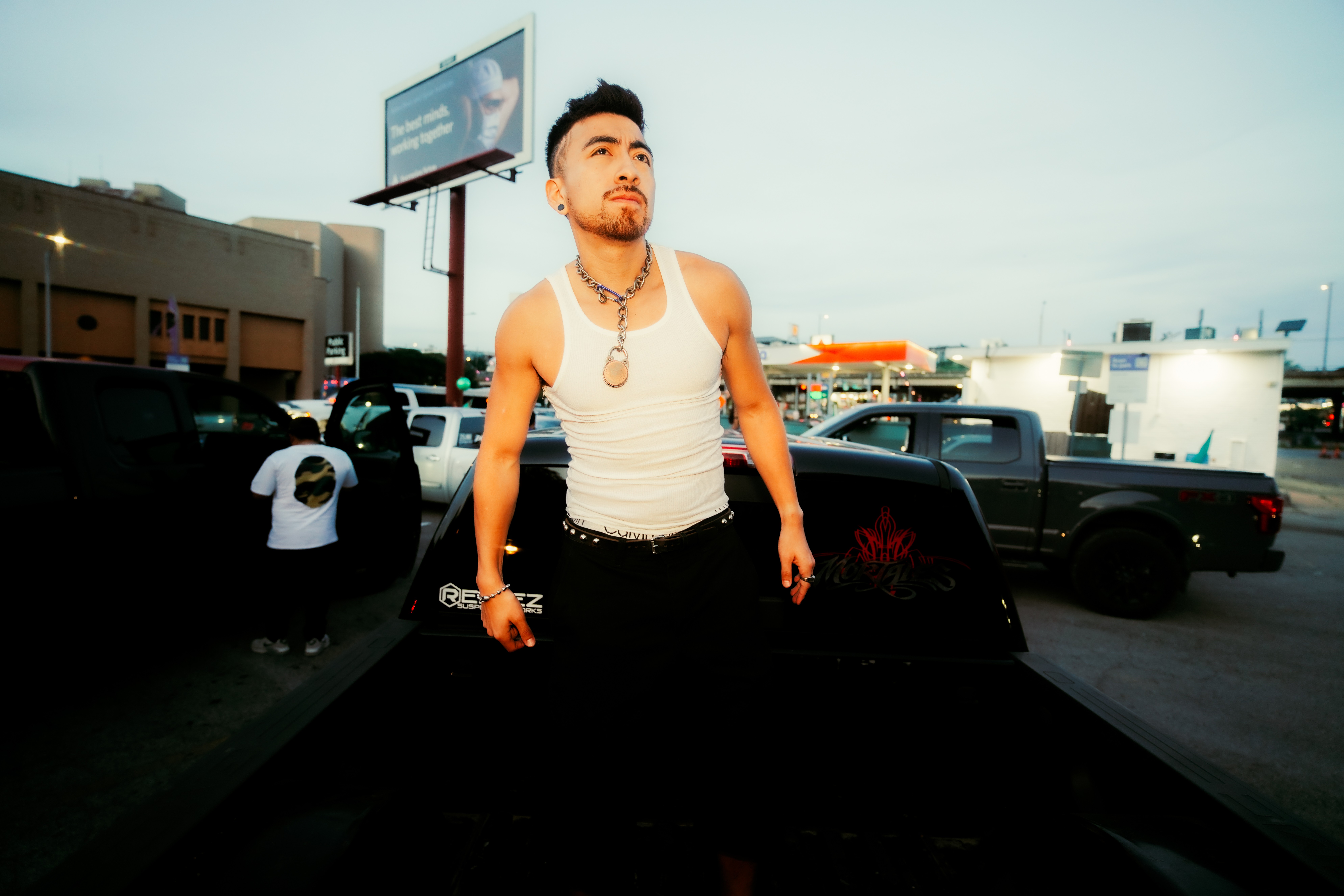 Young man with a necklace and sleeveless shirt standing outdoors near parked cars and a billboard.
