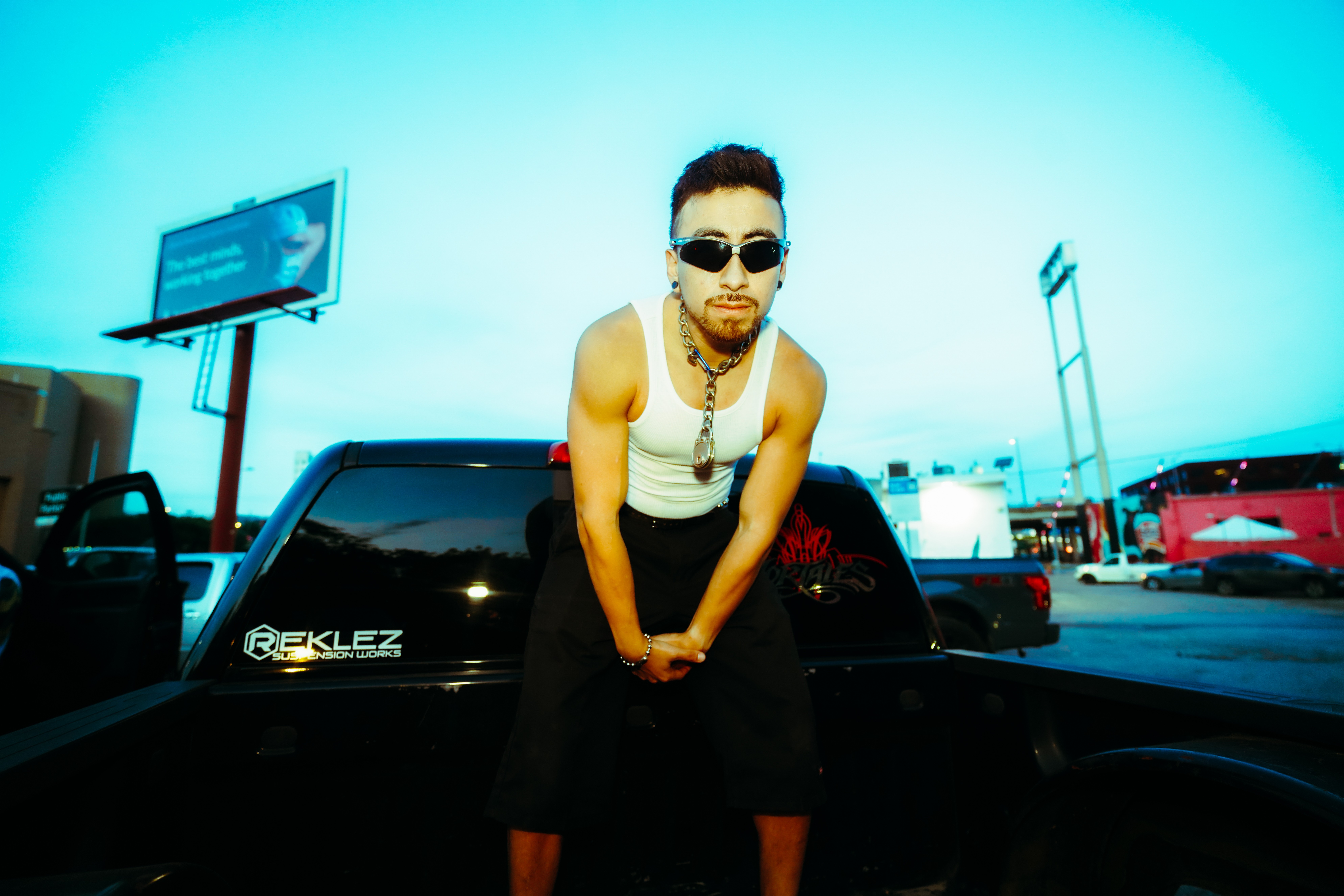 Person with sunglasses and jewelry leaning on a black vehicle in a parking lot, with a blue sky and trucks in background.