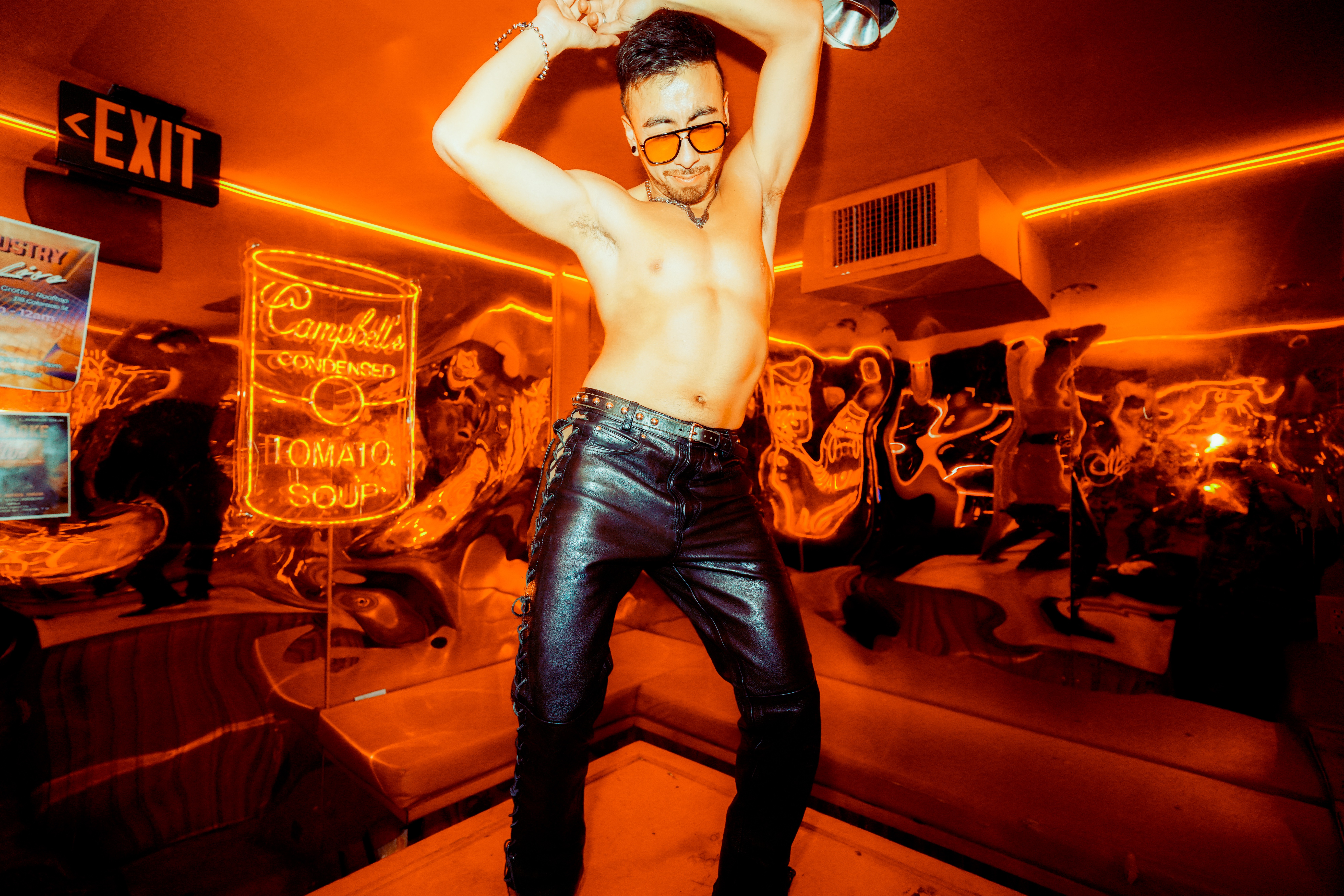 Person with glasses and short hair, shirtless, wearing jeans, dancing in a neon-lit room with graffiti and signs.