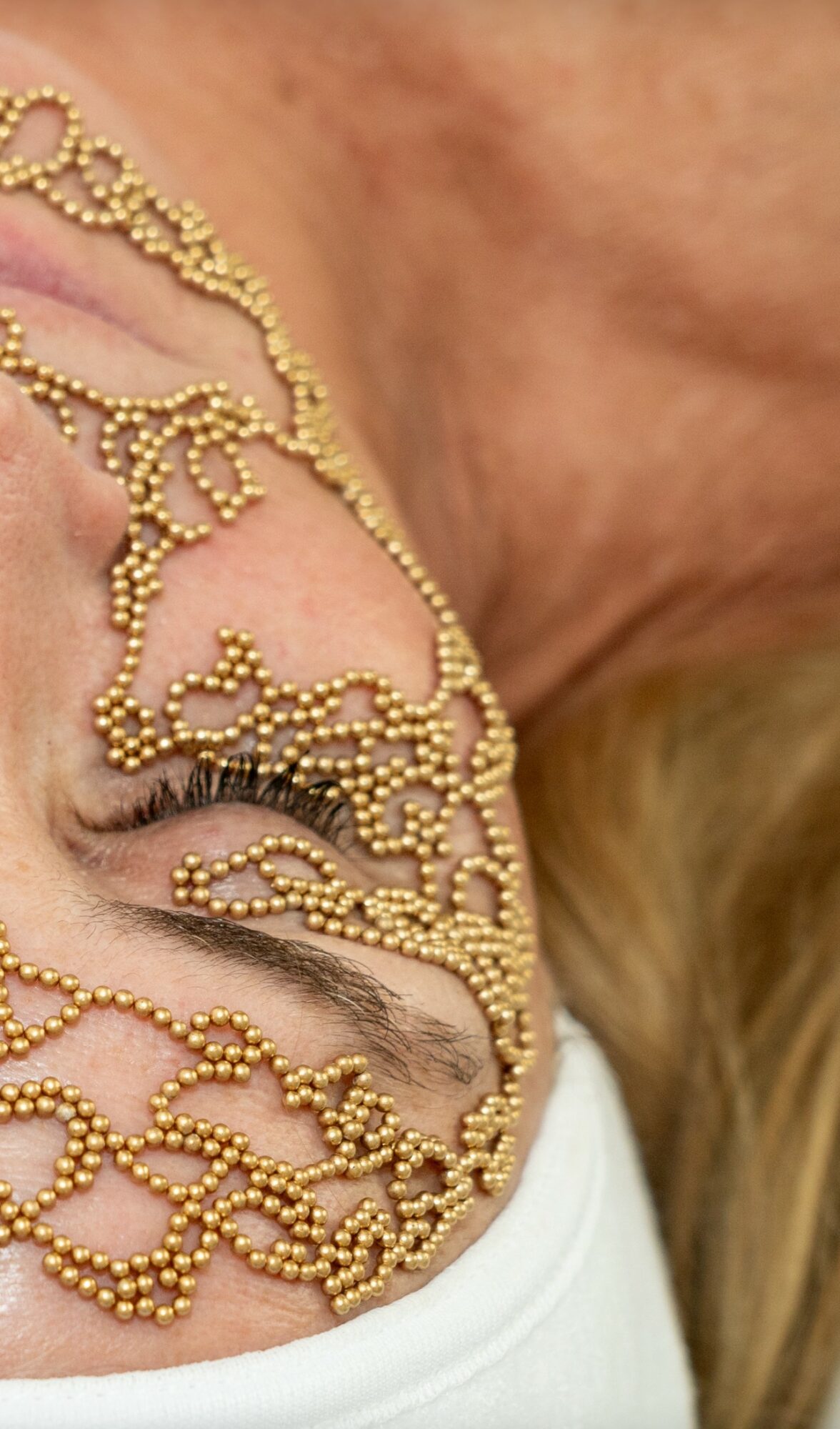 Close-up of a person's face with gold decorative beads covering part of the face and eye, skin visible, light background.