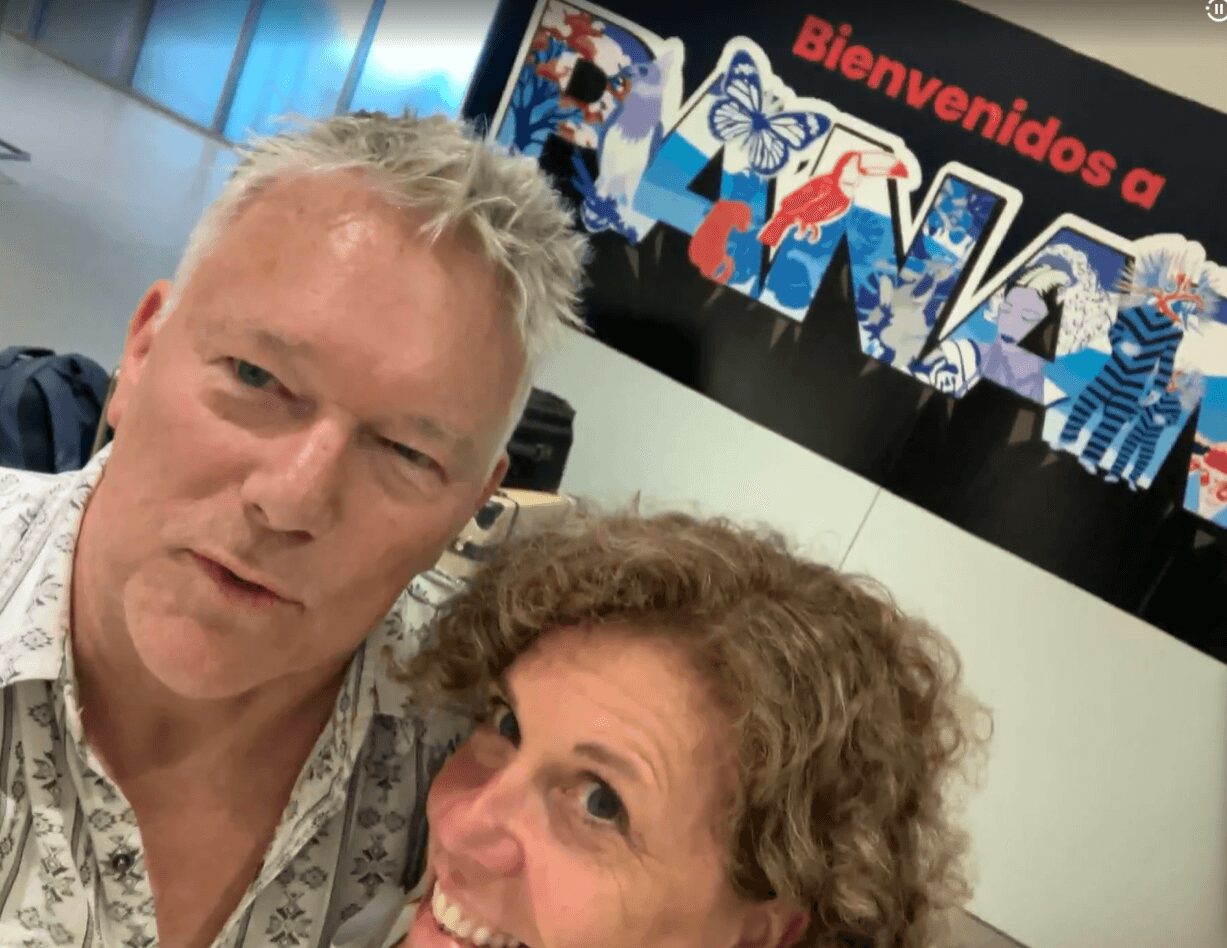 Two people taking a selfie in front of a colorful sign that says 'Bienvenidos a' with decorative images.