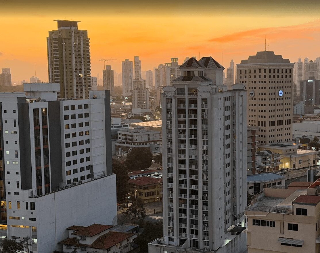 City skyline with tall buildings during sunset, orange sky, and distant high-rise structures.
