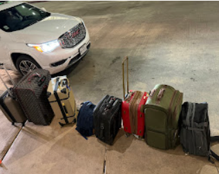 Luggage and backpacks lined up on the sidewalk near a parked white vehicle, with a street and pavement visible in the background.