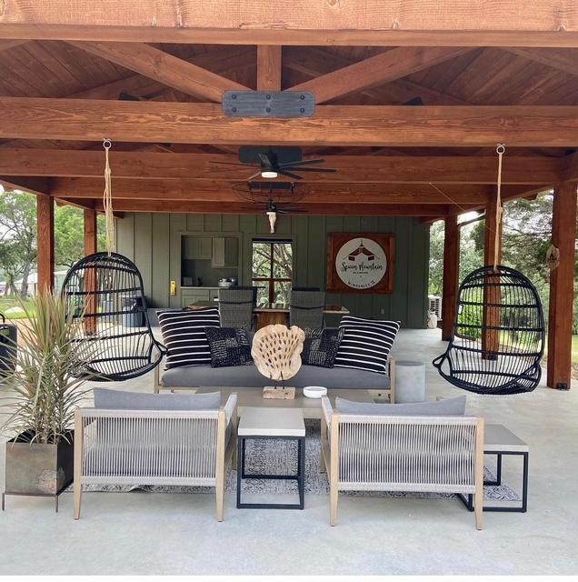 Outdoor seating area with hanging chairs, a sofa, and armchairs under a wooden roof, with a building and trees in background.