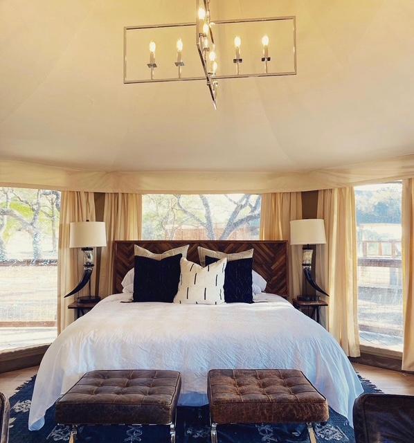 A bedroom with a large bed, pillows, bedside lamps, and windows with curtains, viewed from the foot of the bed.