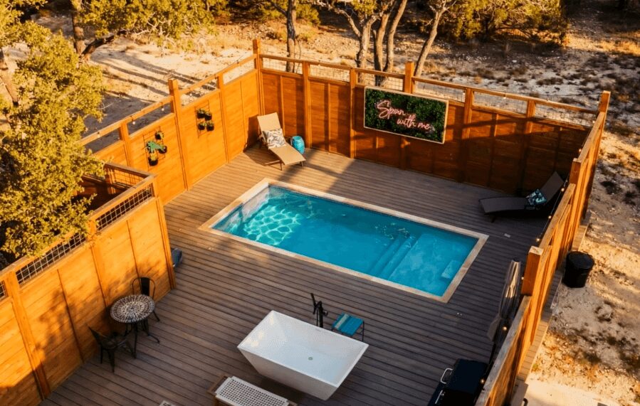 A backyard deck with a swimming pool, lounge chairs, a table, and a chalkboard, surrounded by a wooden fence and trees.