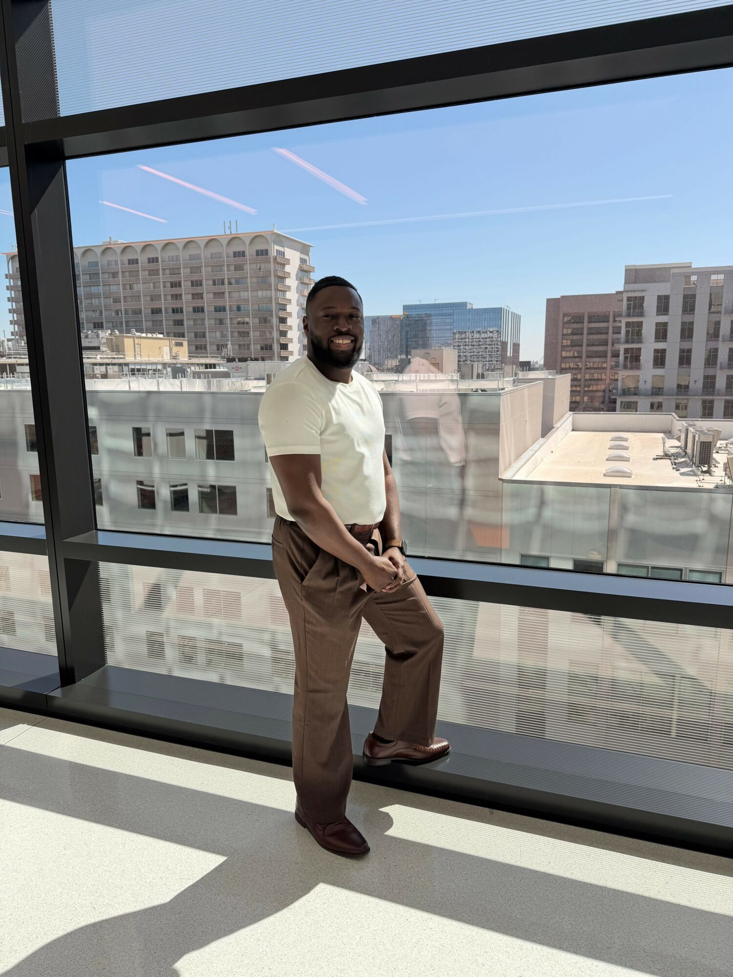 Man standing indoors near large window with cityscape view, wearing white shirt and brown pants, looking at camera.