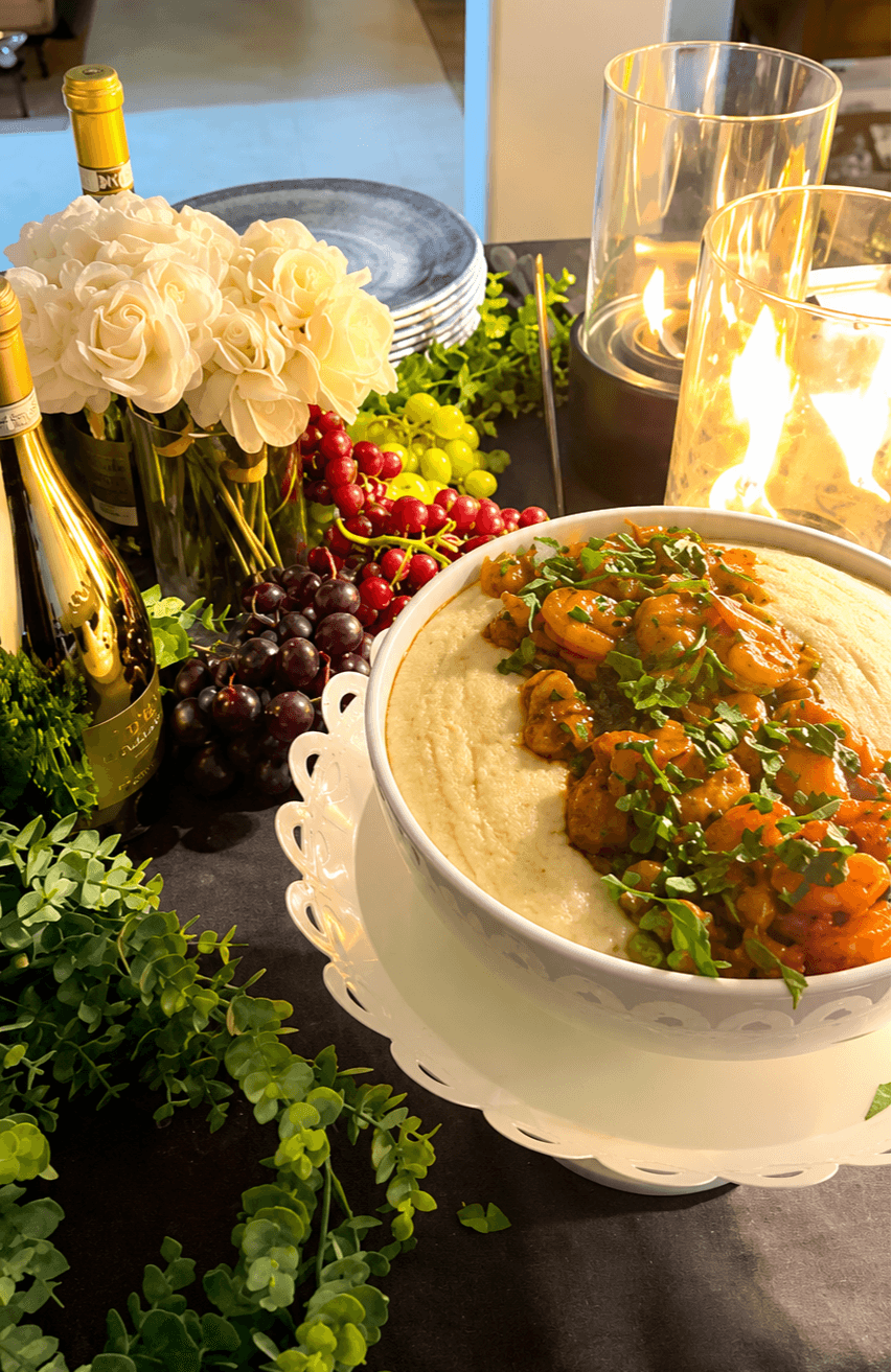 Table with a bowl of stew, flowers, grapes, champagne, candles, and greenery, set for a meal, in warm lighting.