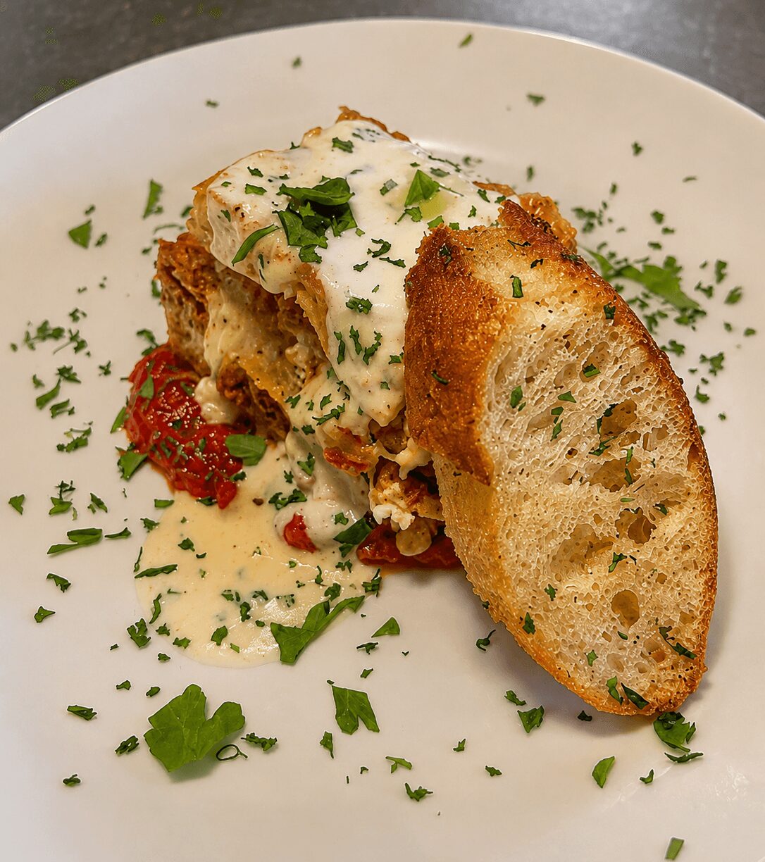 Slice of bread with filling, topped with sauce and herbs, on a white plate with garnishes.