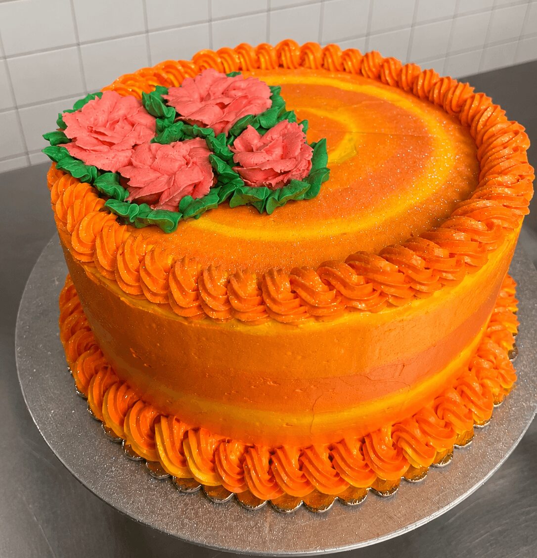 Round cake with orange and yellow frosting, pink flowers with green leaves decoration, on a silver base.