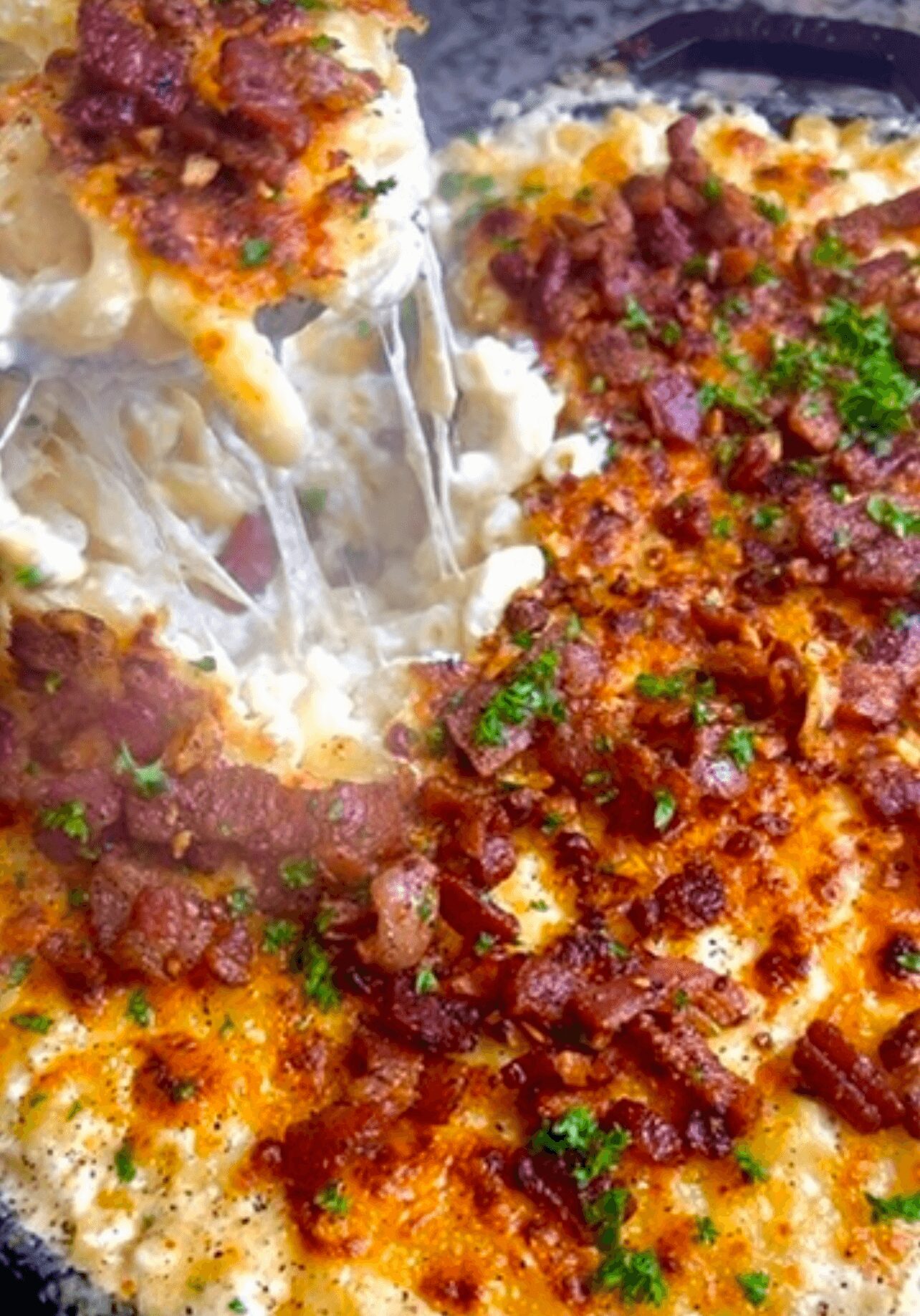 Close-up of baked cheesy dish topped with bacon bits and chopped herbs, with a spoon lifting a portion, showing melted cheese.