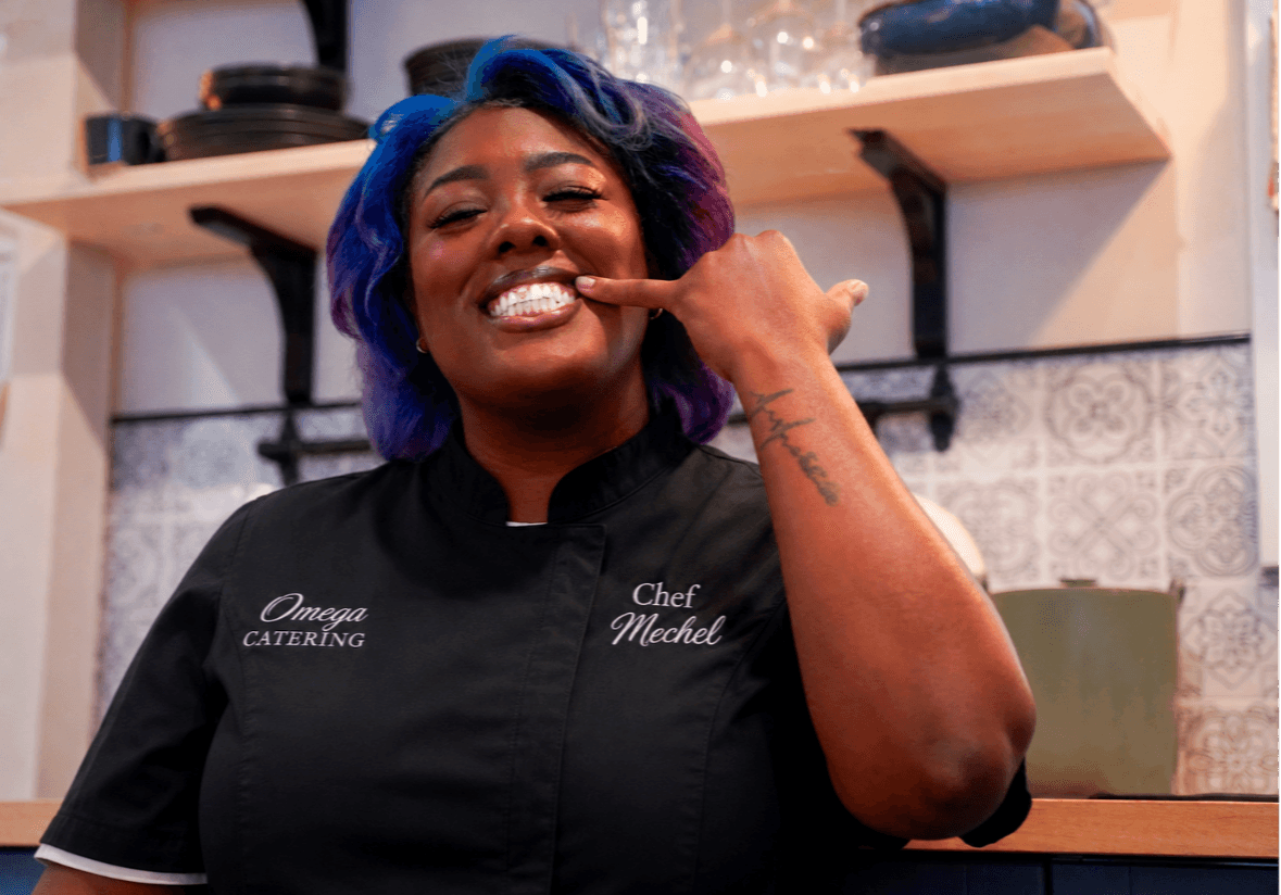 Woman with colorful hair smiling and making a hand gesture in a kitchen setting.