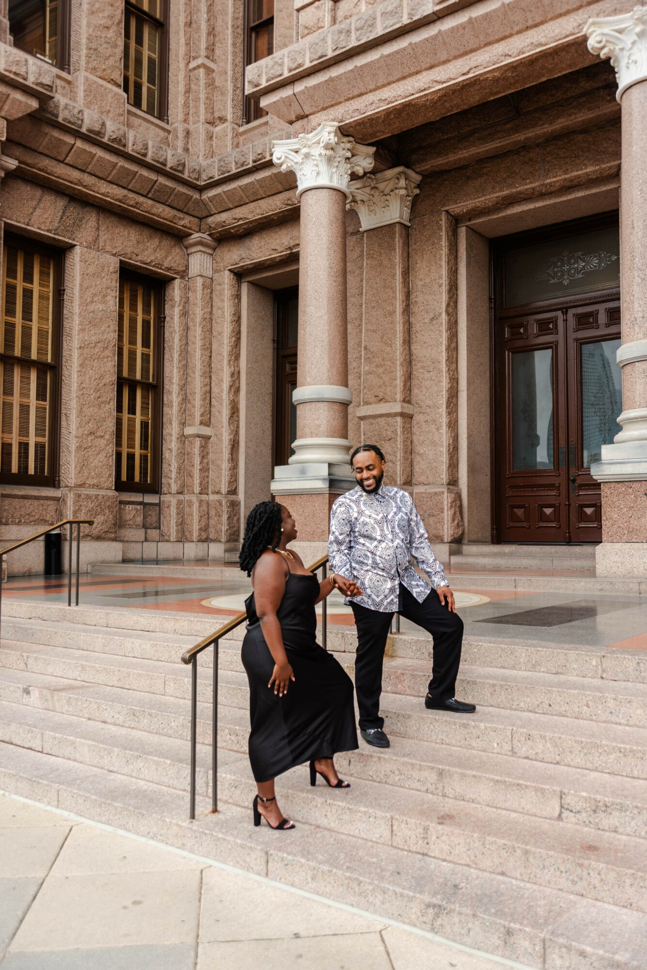 Two people standing on steps outside a grand building with columns and large doors, engaged in conversation.