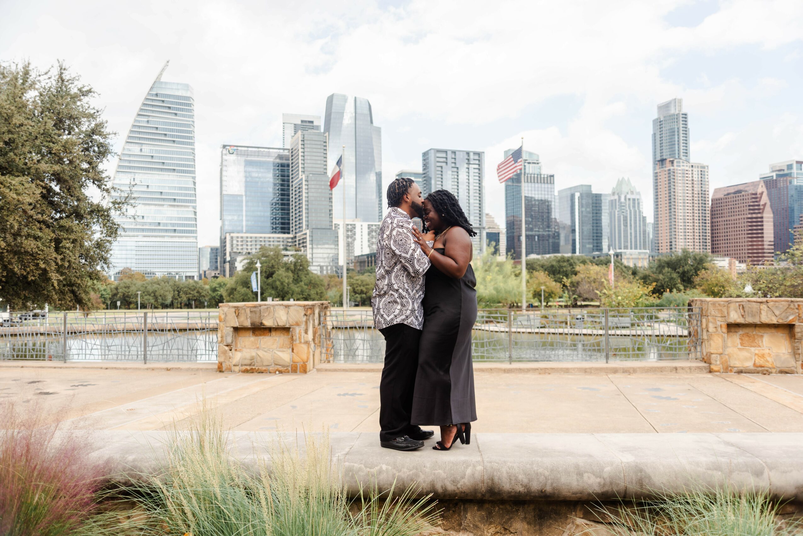 Two people embrace on a waterfront promenade with a city skyline in the background, trees, and a cloudy sky.