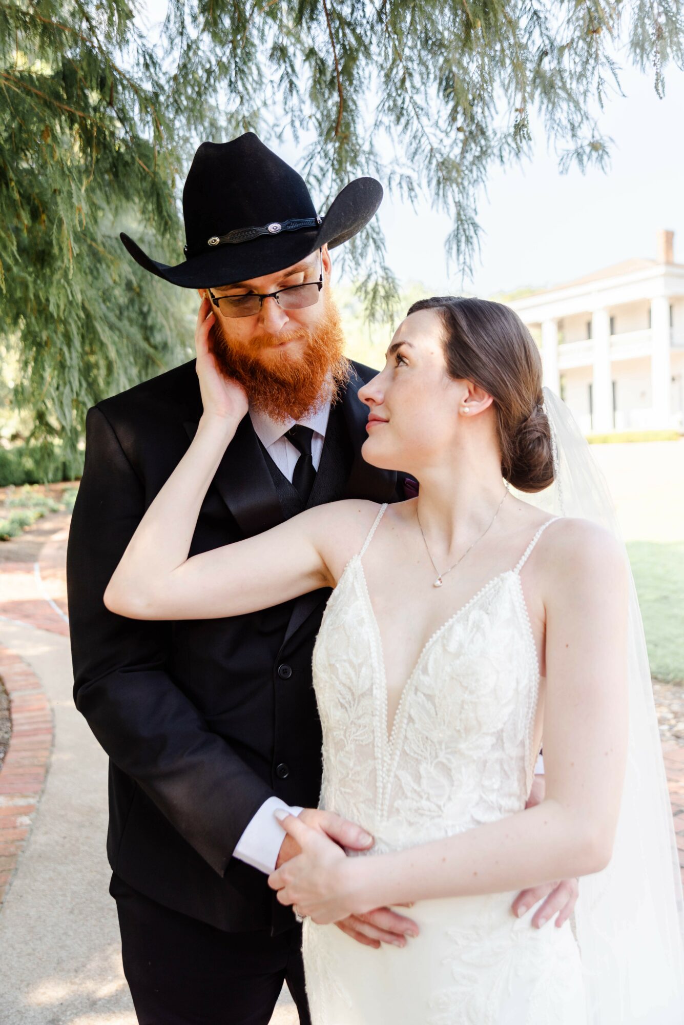 A man with a beard and glasses wearing a black hat and suit, and a woman in a white dress, standing outdoors under trees.