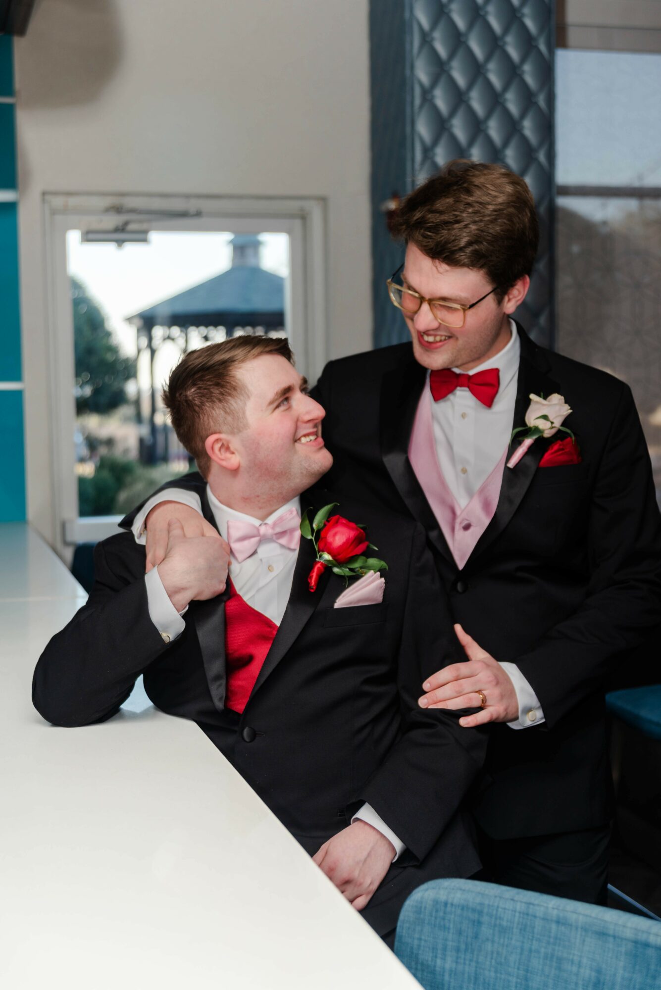Two young men in tuxedos with pink vests and red bow ties smiling and looking at each other, one with glasses, indoors.
