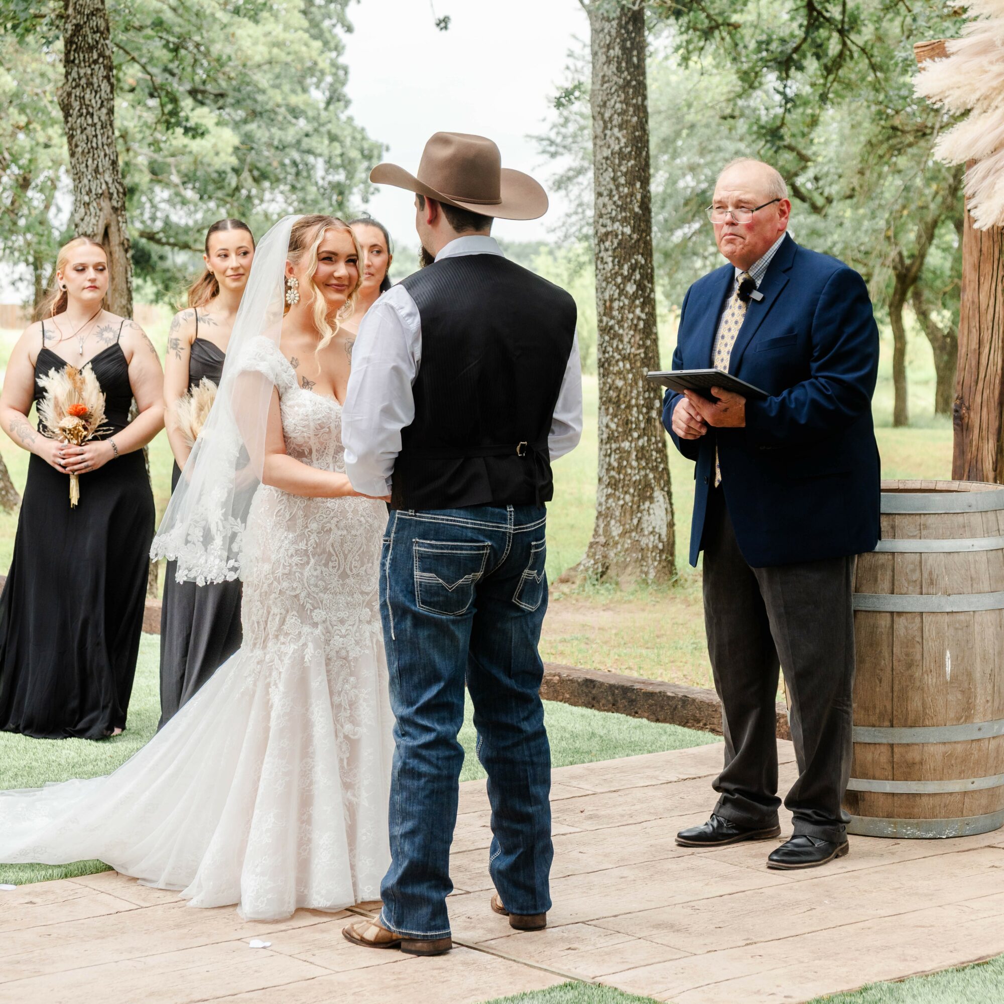 Bride and groom exchanging vows outdoors, officiant holding a book, bridesmaids in black dresses holding bouquets, trees in background.