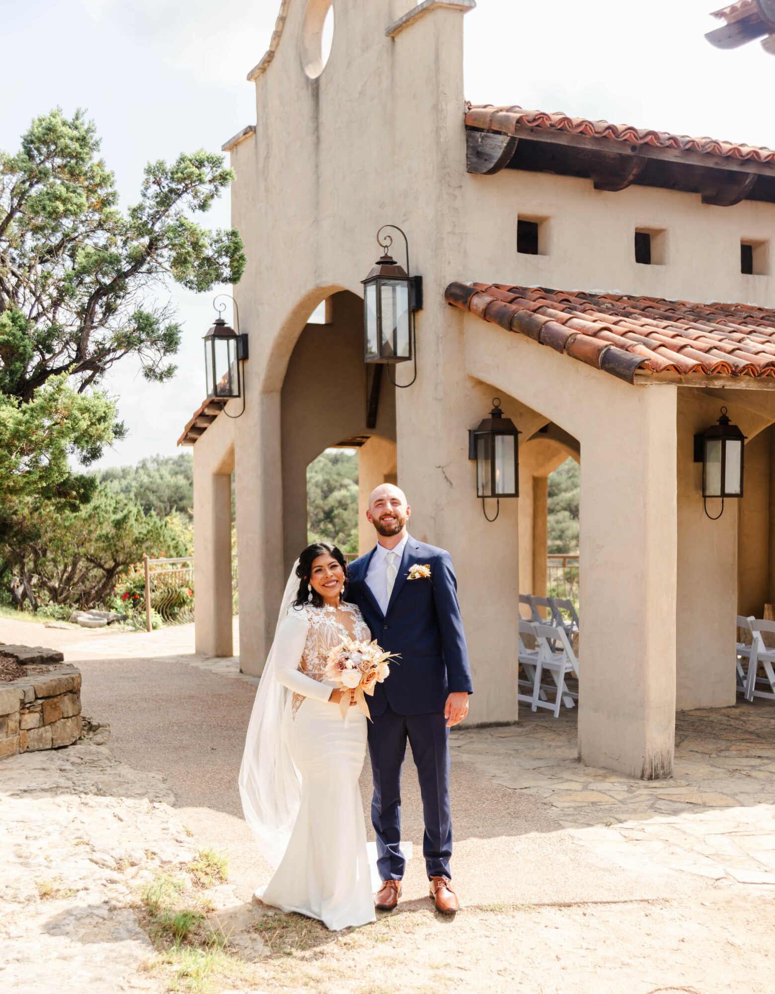 Bride and groom standing outside a building with arches and lanterns, smiling, holding flowers, sunny day, trees in background.