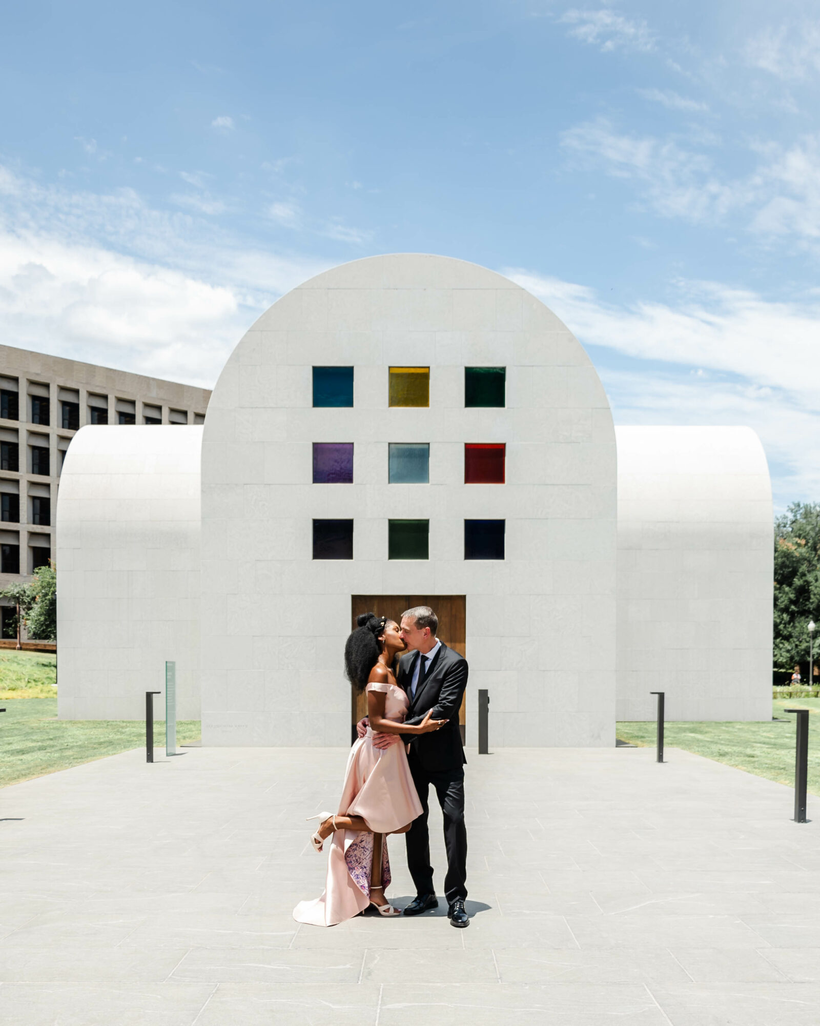 Couple embracing in front of a modern white building with colorful square windows, under a partly cloudy sky.
