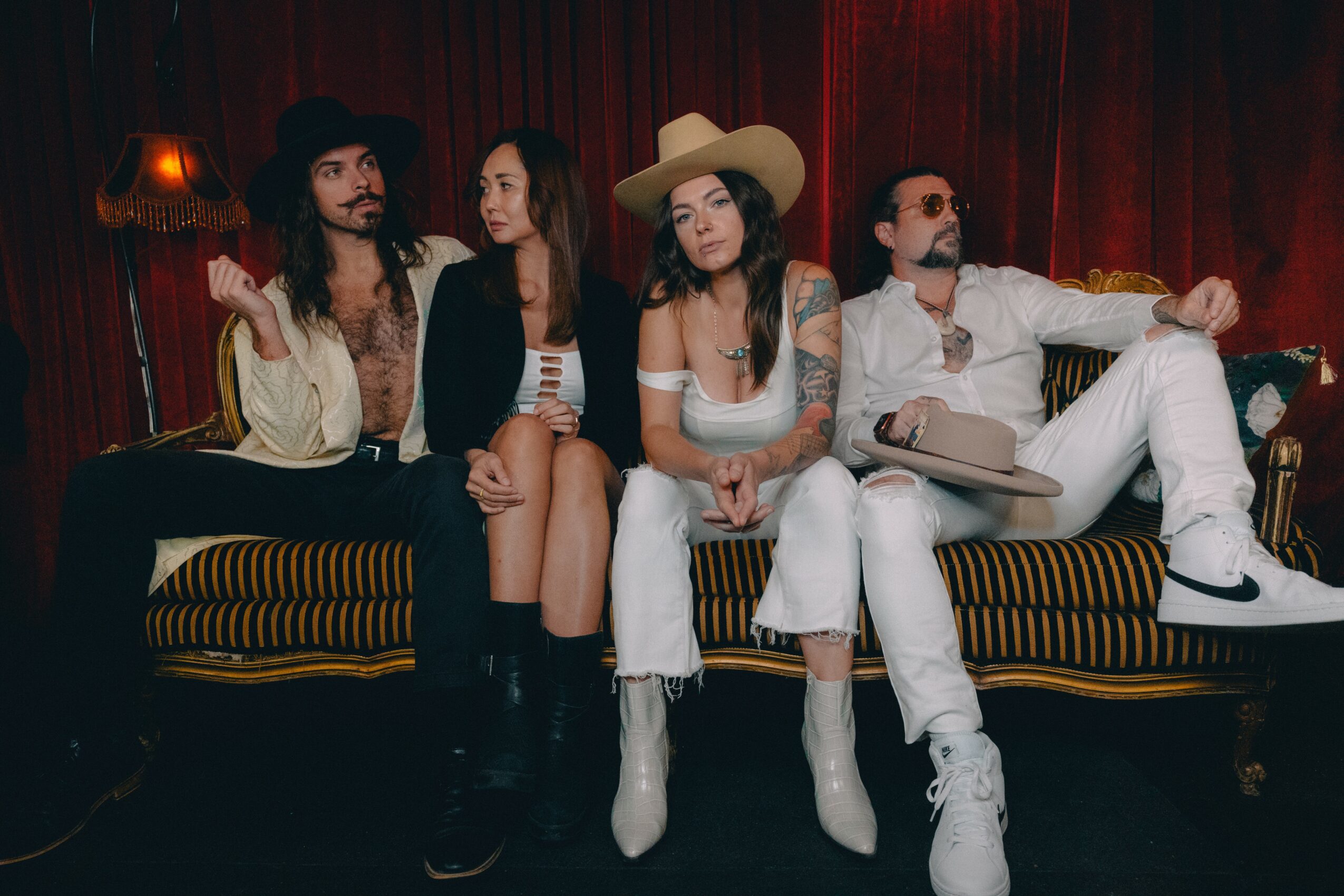 Four people sitting on a striped sofa in front of a red curtain, dressed in vintage and modern clothing, some with hats.
