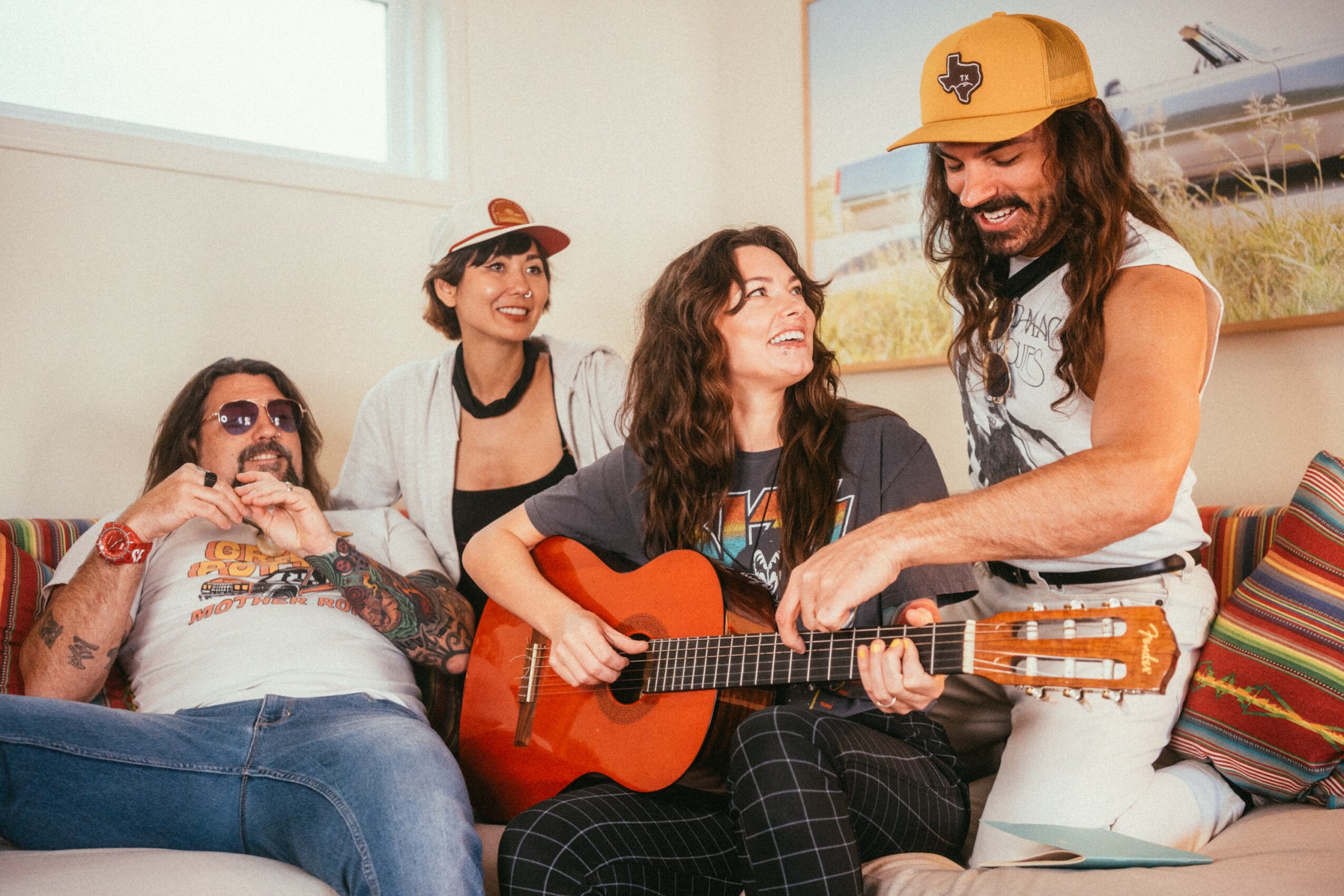 Four people in a room, one playing guitar, others smiling and sitting on a couch with colorful pillows.