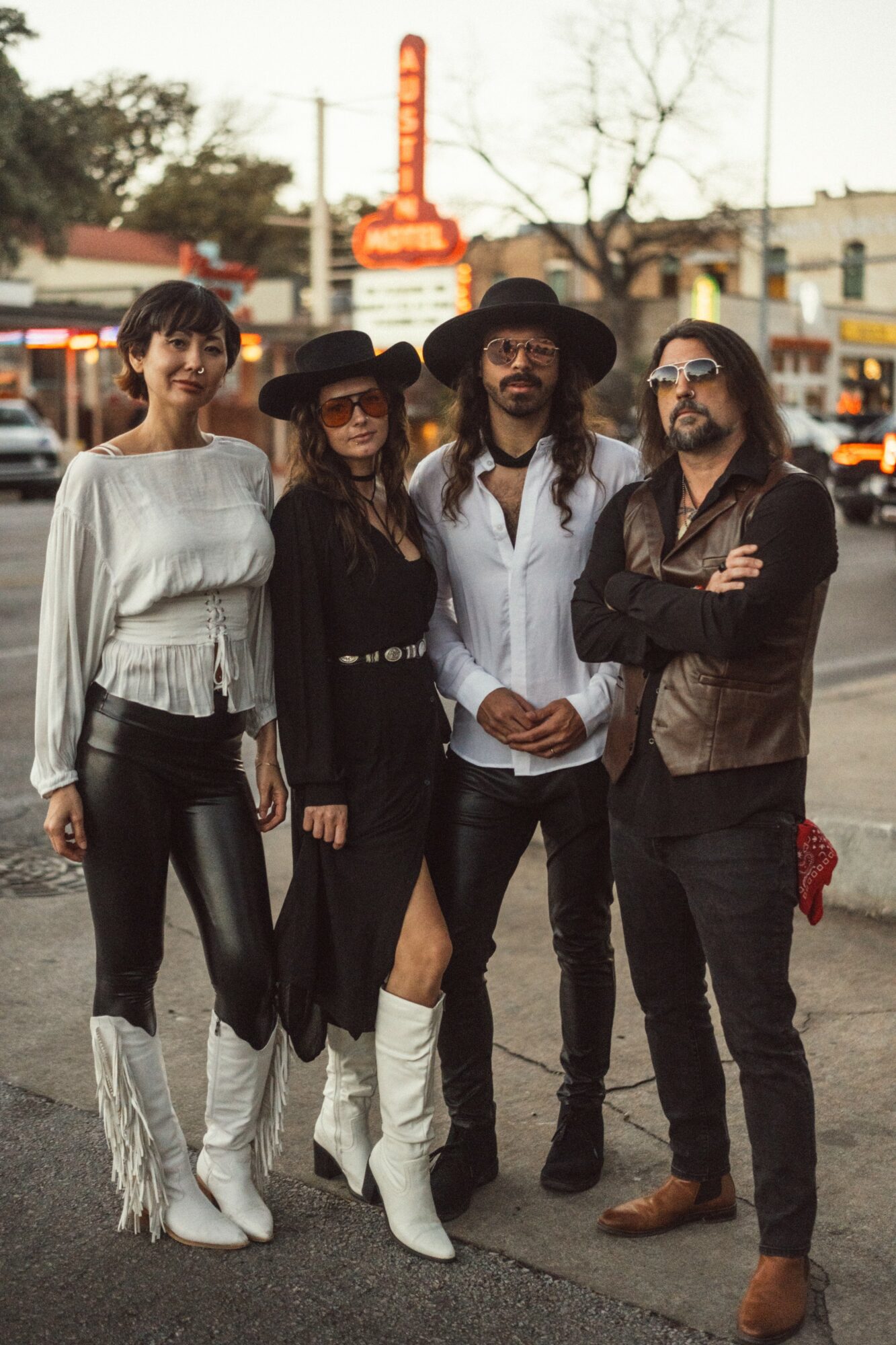 Four people standing outdoors on a street, dressed in stylish clothing, with buildings and trees in the background.