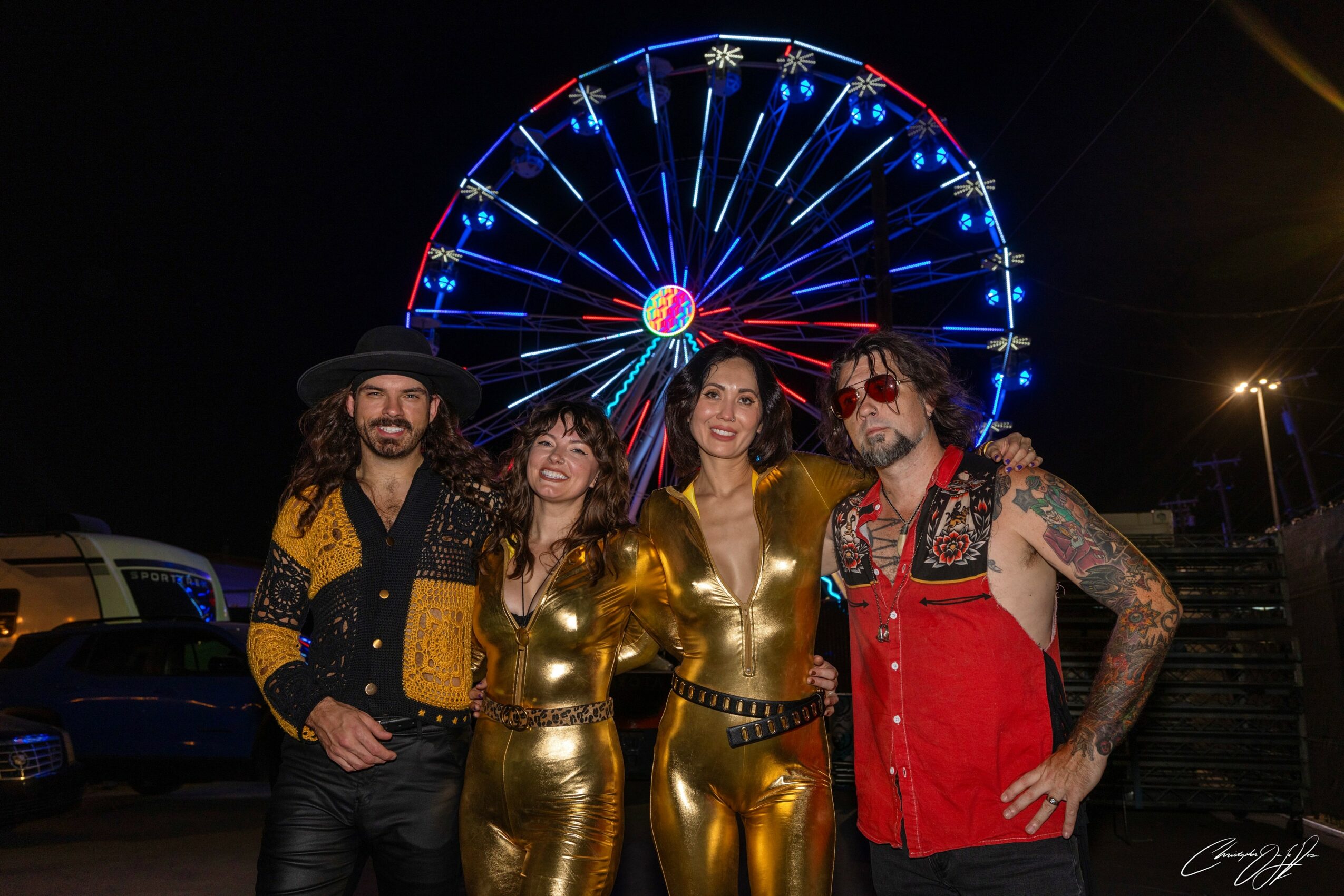 Four people in costumes stand in front of a brightly lit Ferris wheel at night, smiling and posing together.