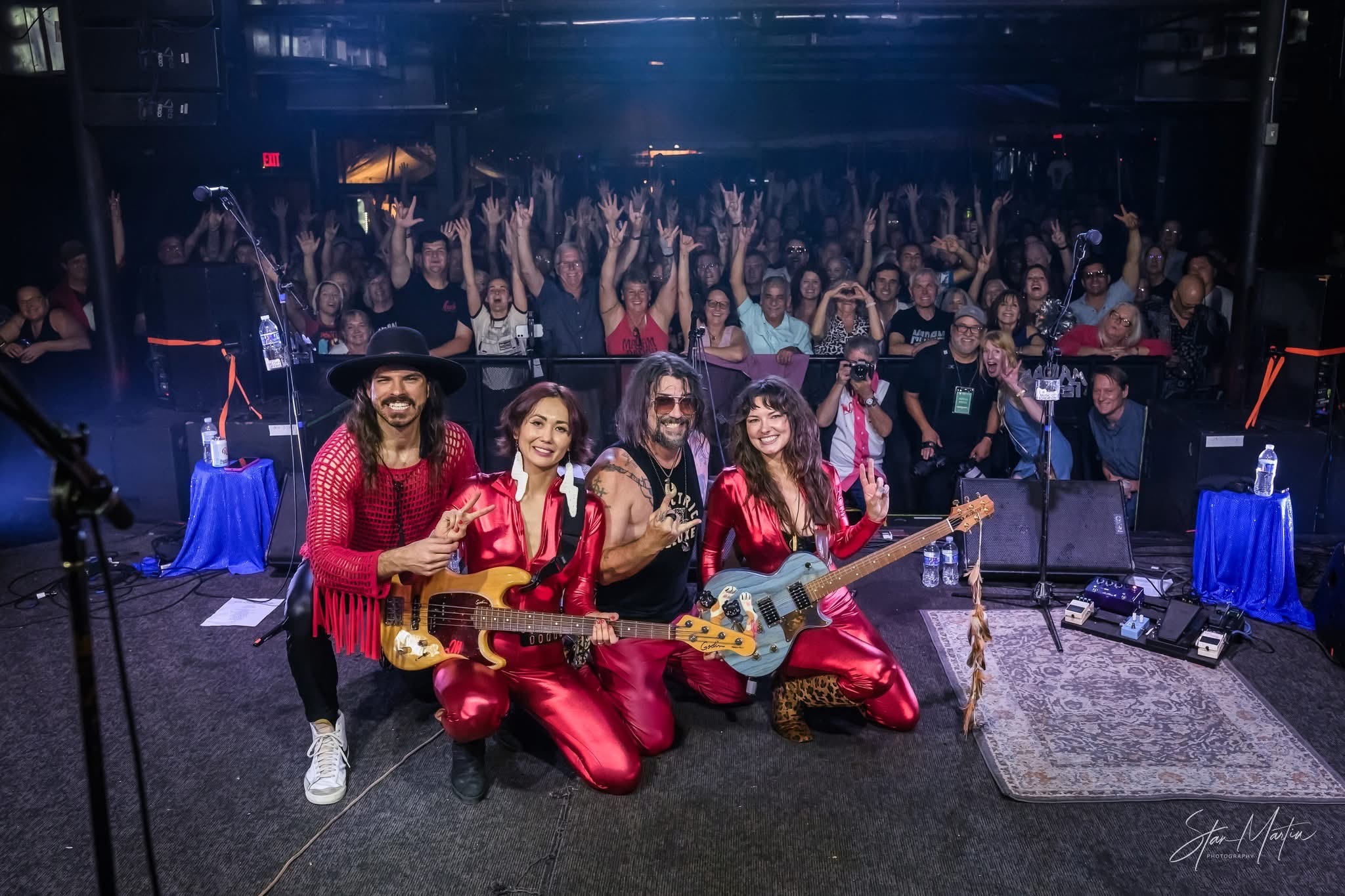 Four musicians in red outfits with guitars perform on stage, audience claps and raises hands in background.