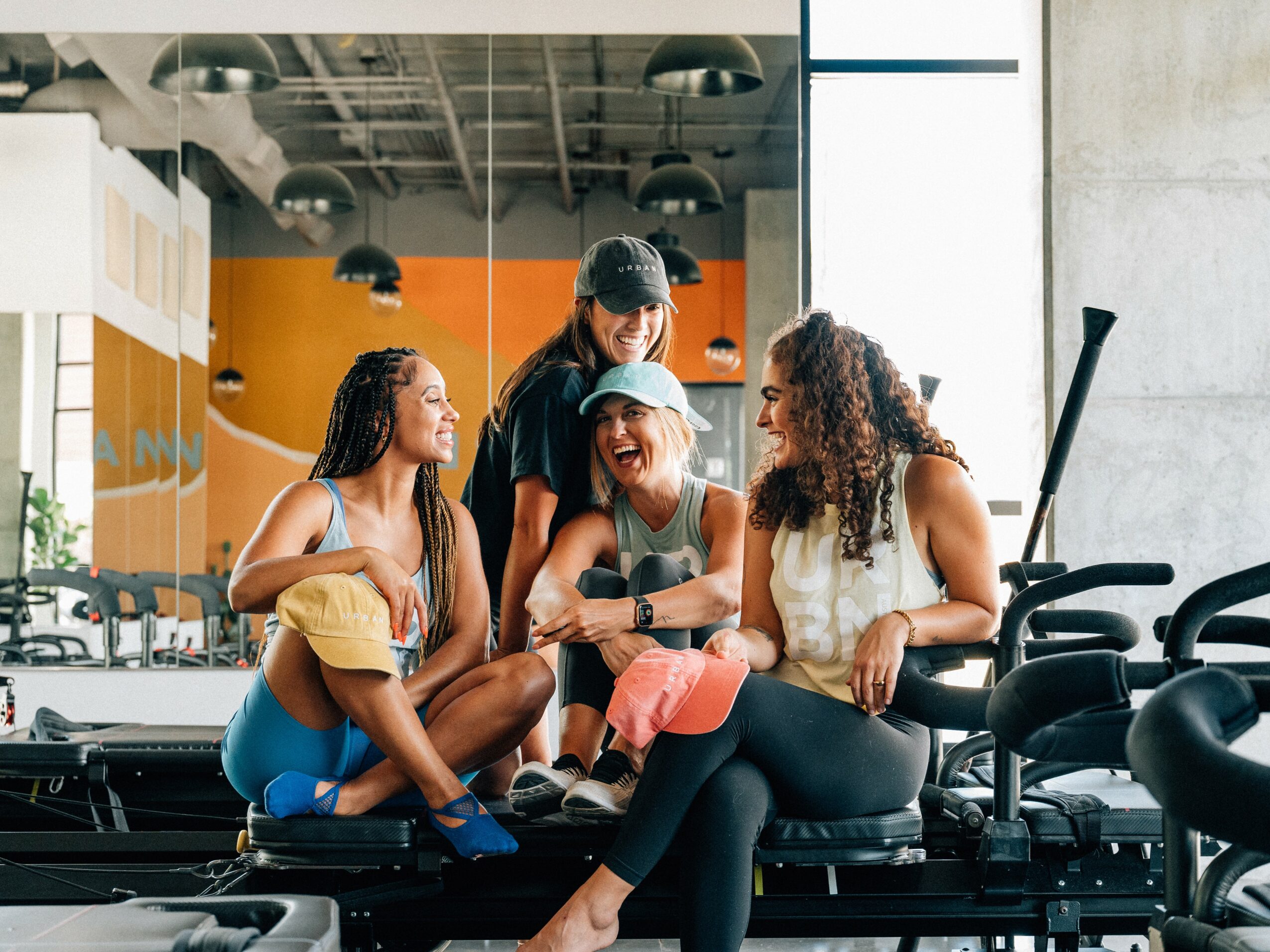 Four people sitting on a gym mat, smiling and talking, with gym equipment in the background.