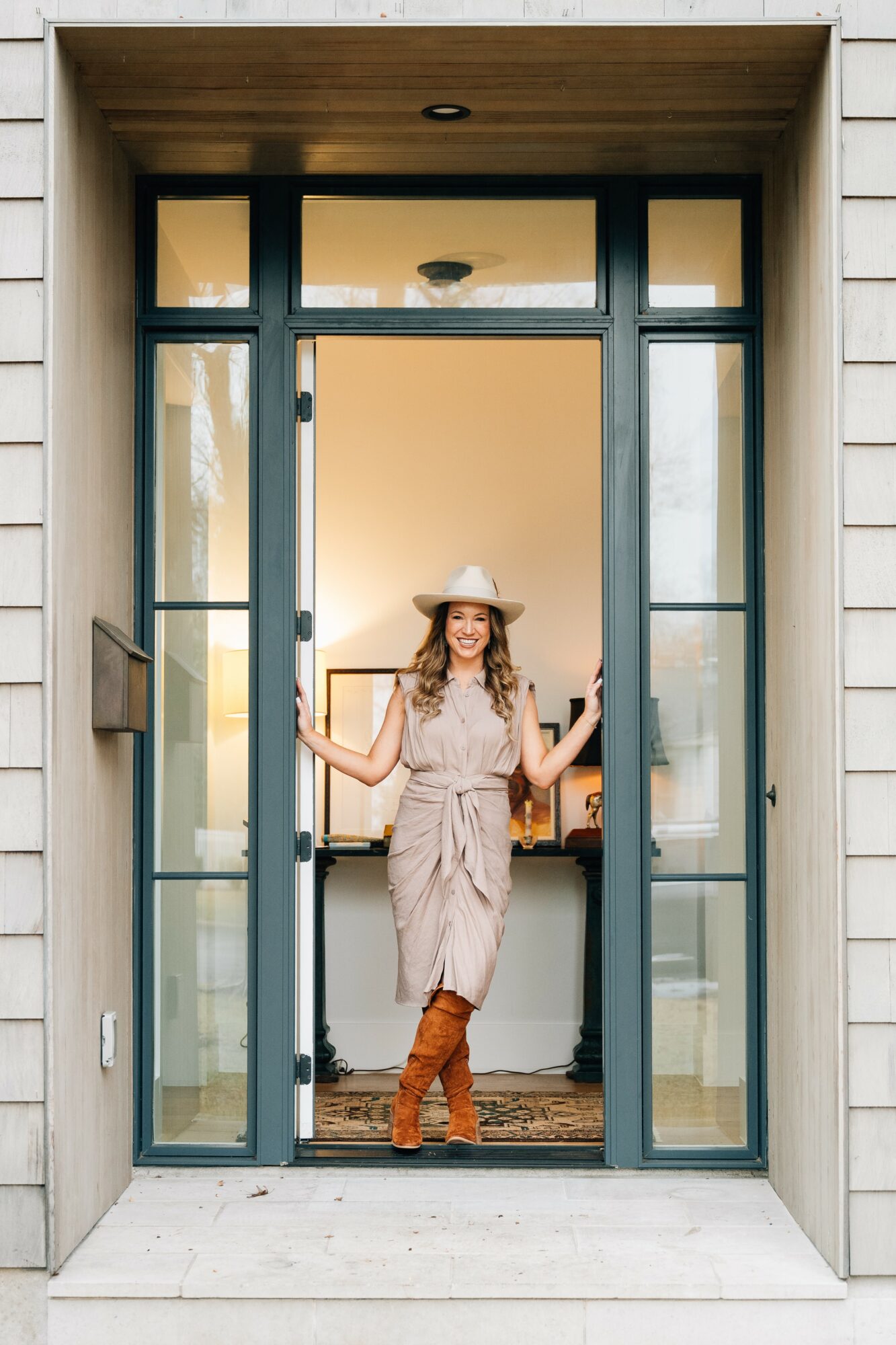 Woman standing in doorway, wearing a hat and boots, smiling, with a cozy interior visible behind her.