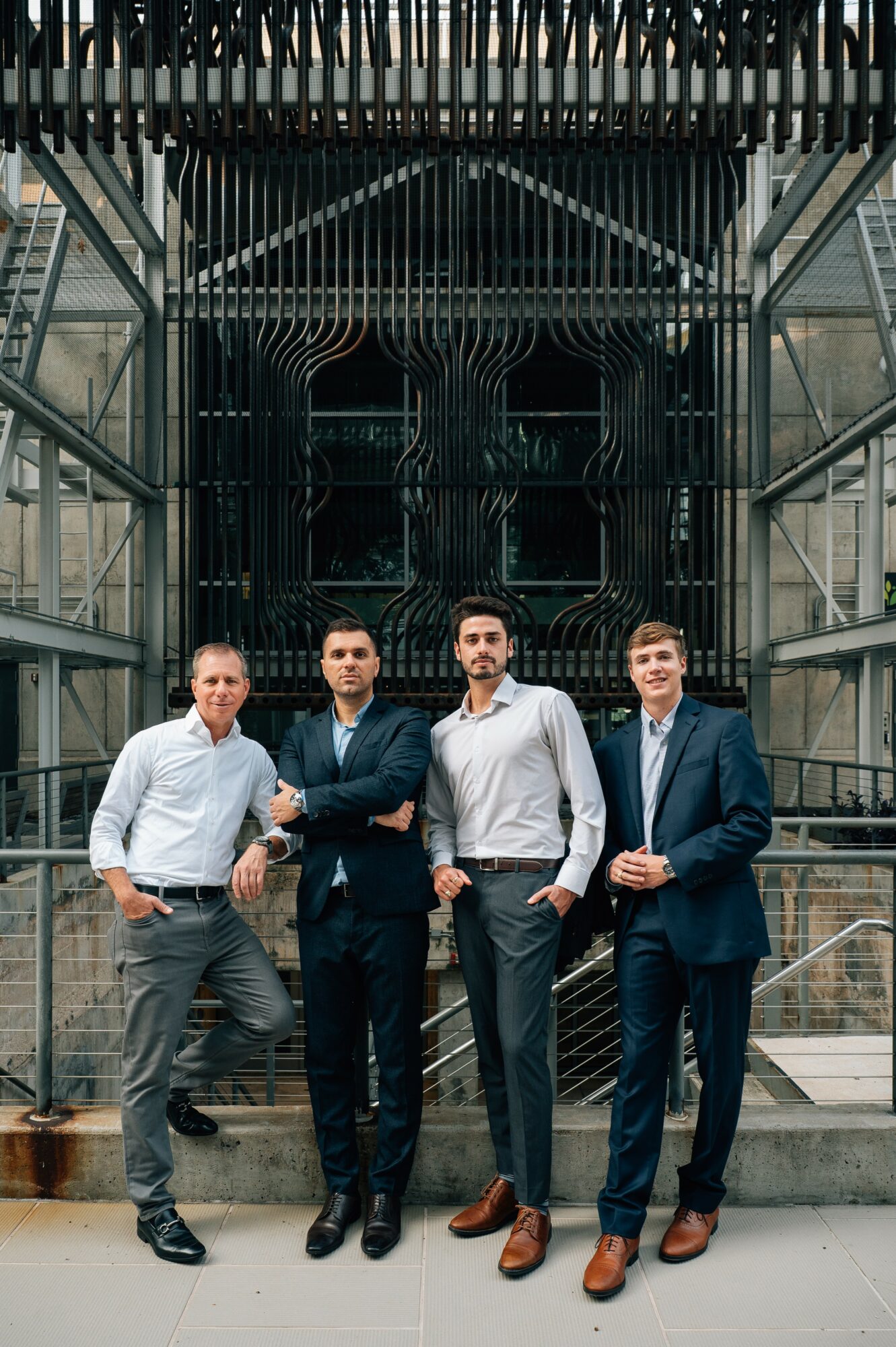 Four men stand in front of a large, ornate black gate with metal framework and glass building behind them.