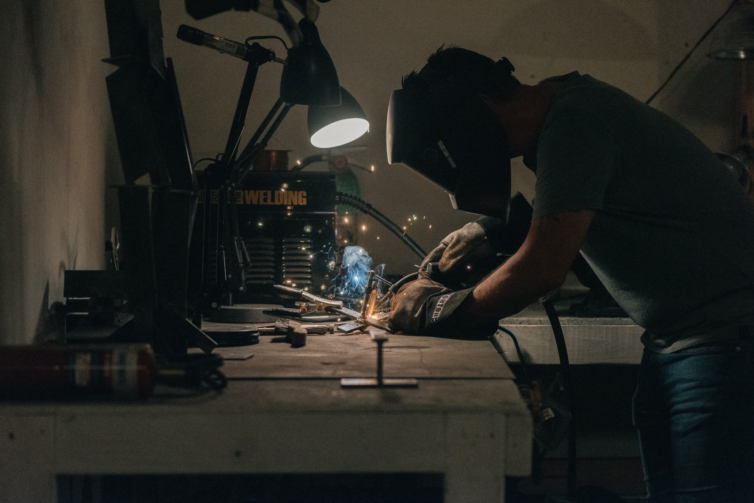 Person welding at a workbench in a dimly lit workshop, sparks flying from welding tool, focused on work.