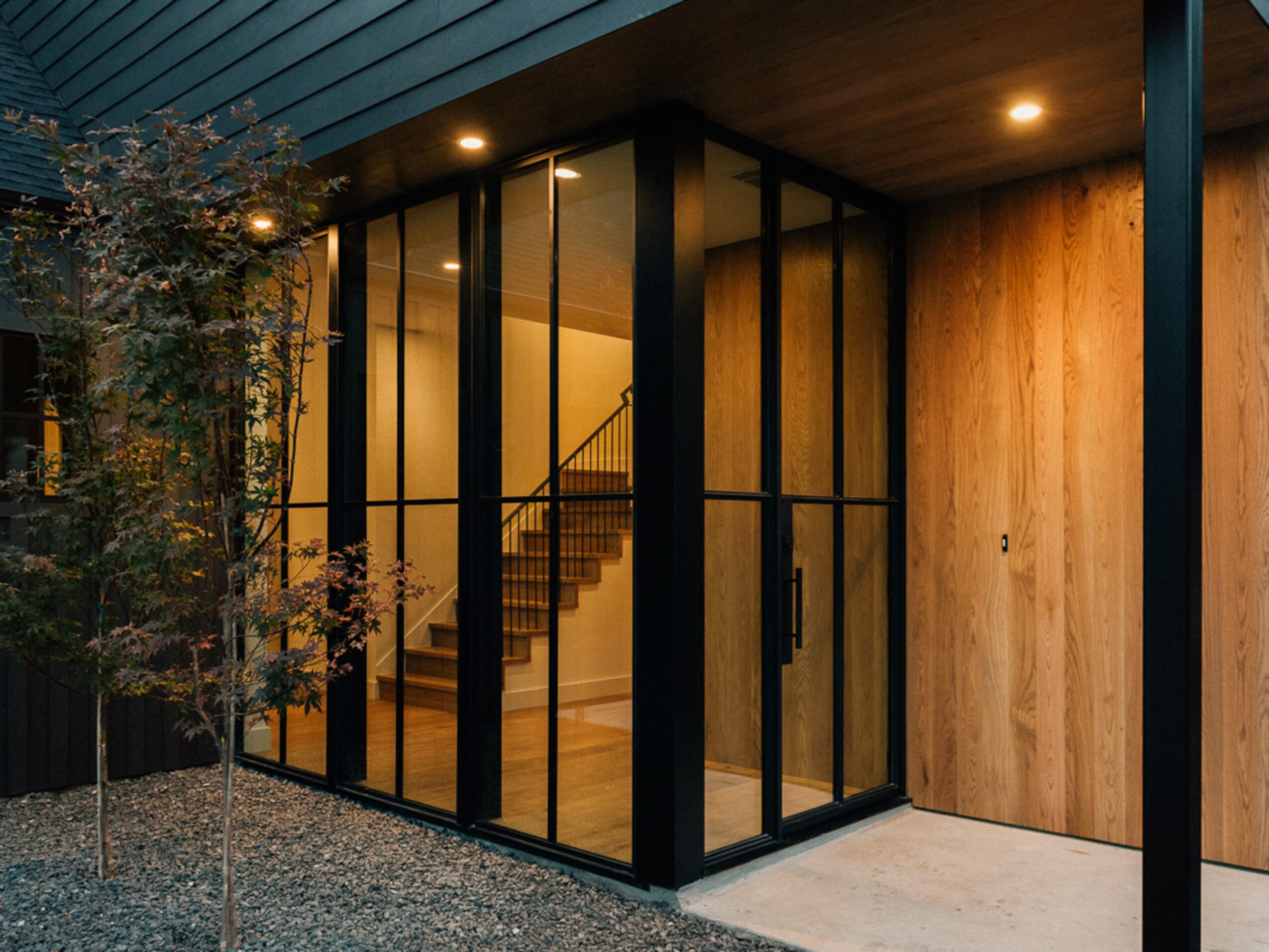 Modern building entrance with glass walls, wooden paneling, and a staircase visible inside, illuminated by ceiling lights.