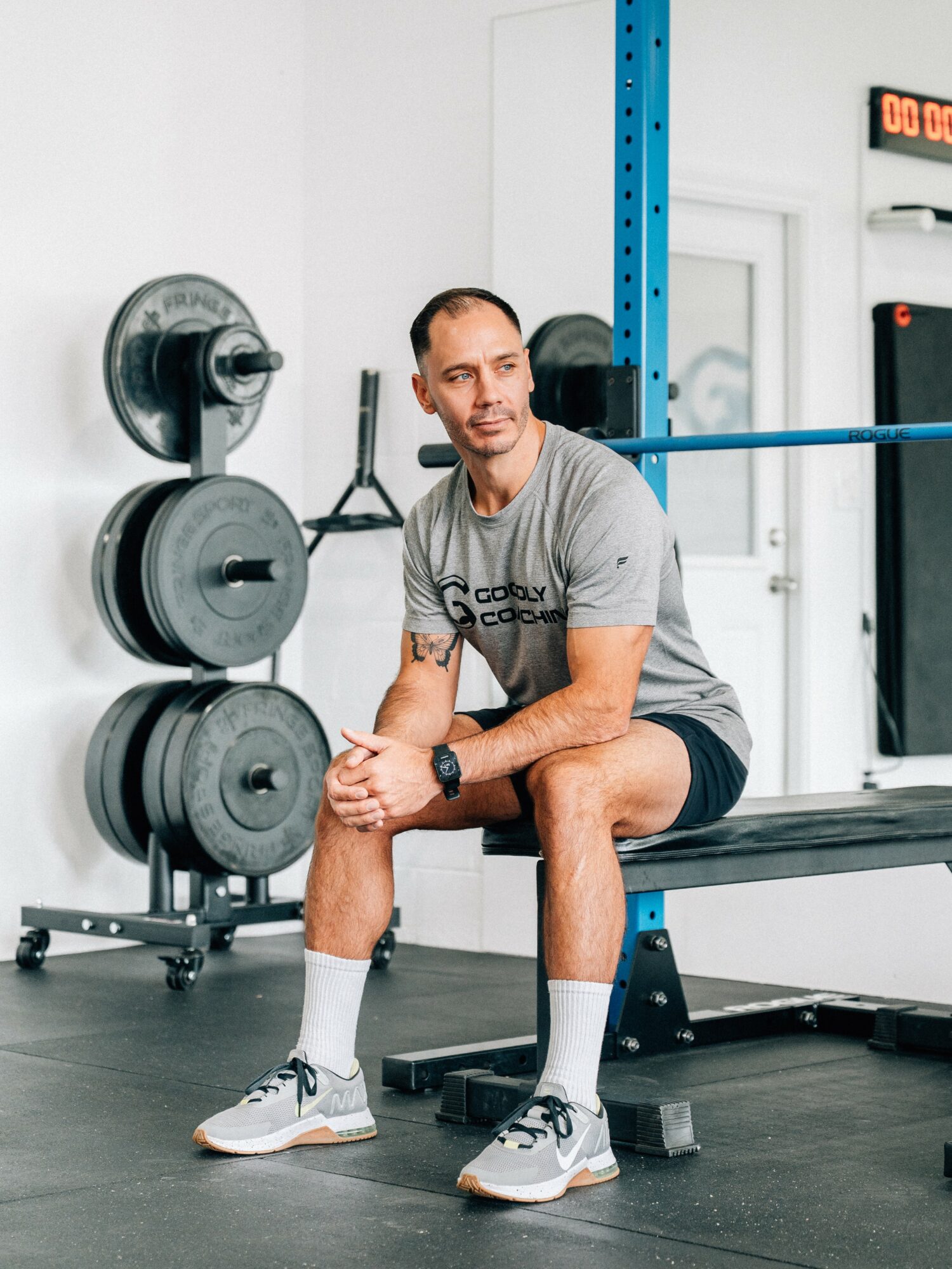 Man sitting on a bench in a gym, with weight plates and equipment behind him.