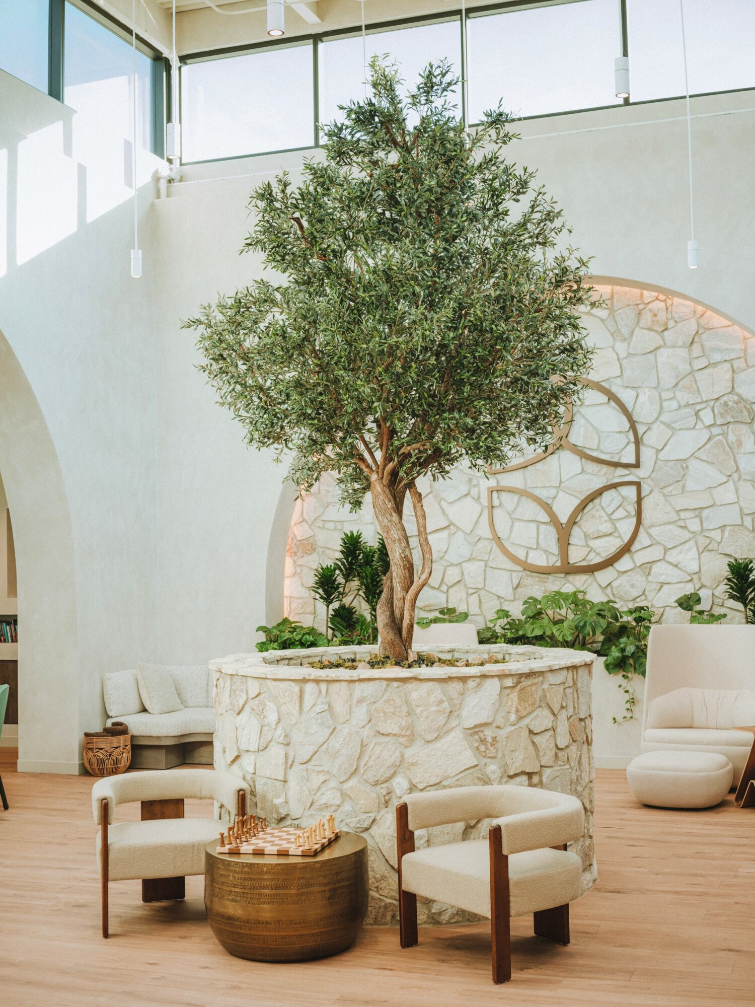 Indoor space with a large tree in a stone planter, surrounded by chairs and a wooden table, with a stone wall and windows.