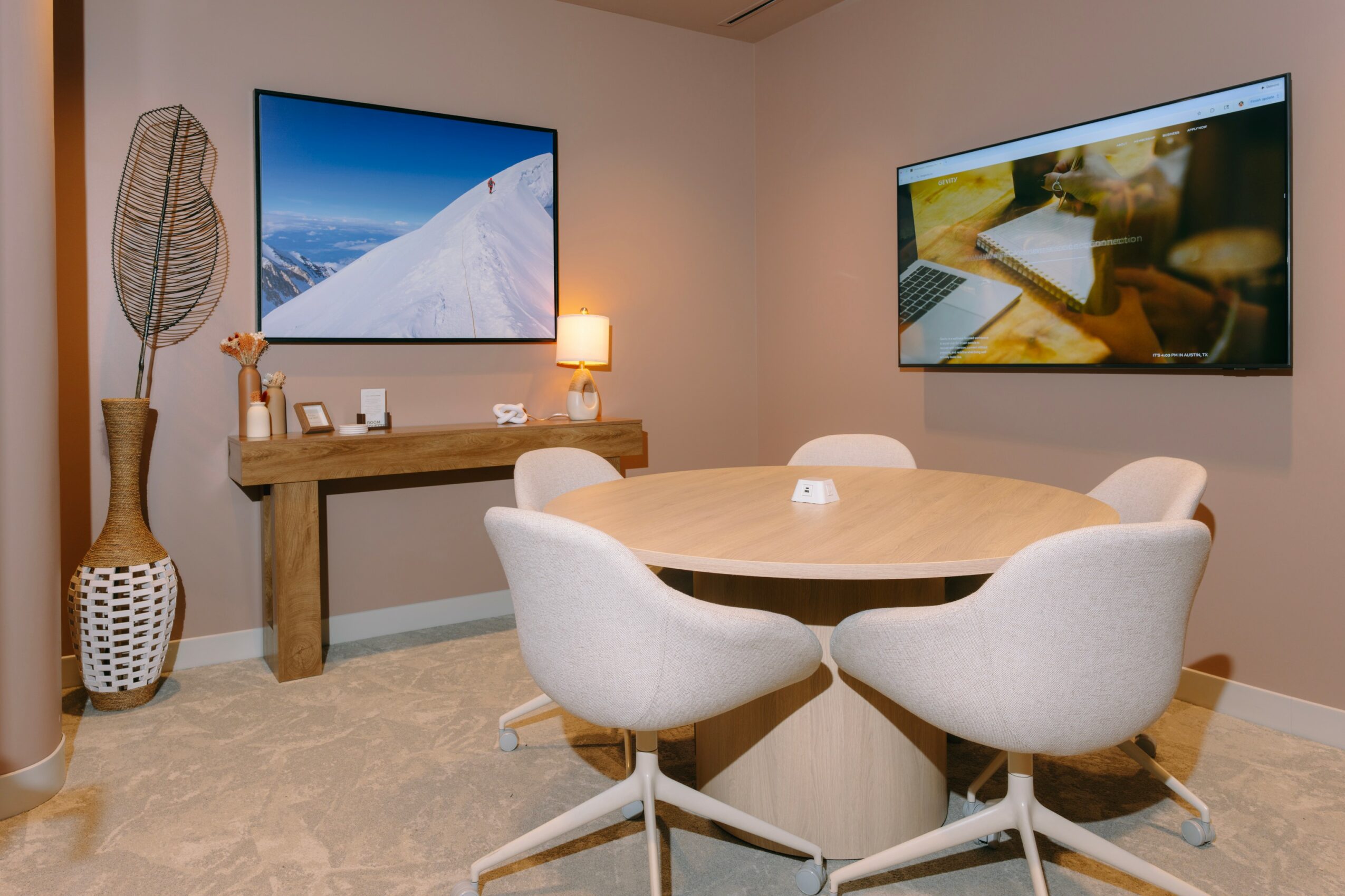 Round table with four white chairs in a room with two wall-mounted screens, a wooden console, and a tall decorative vase.