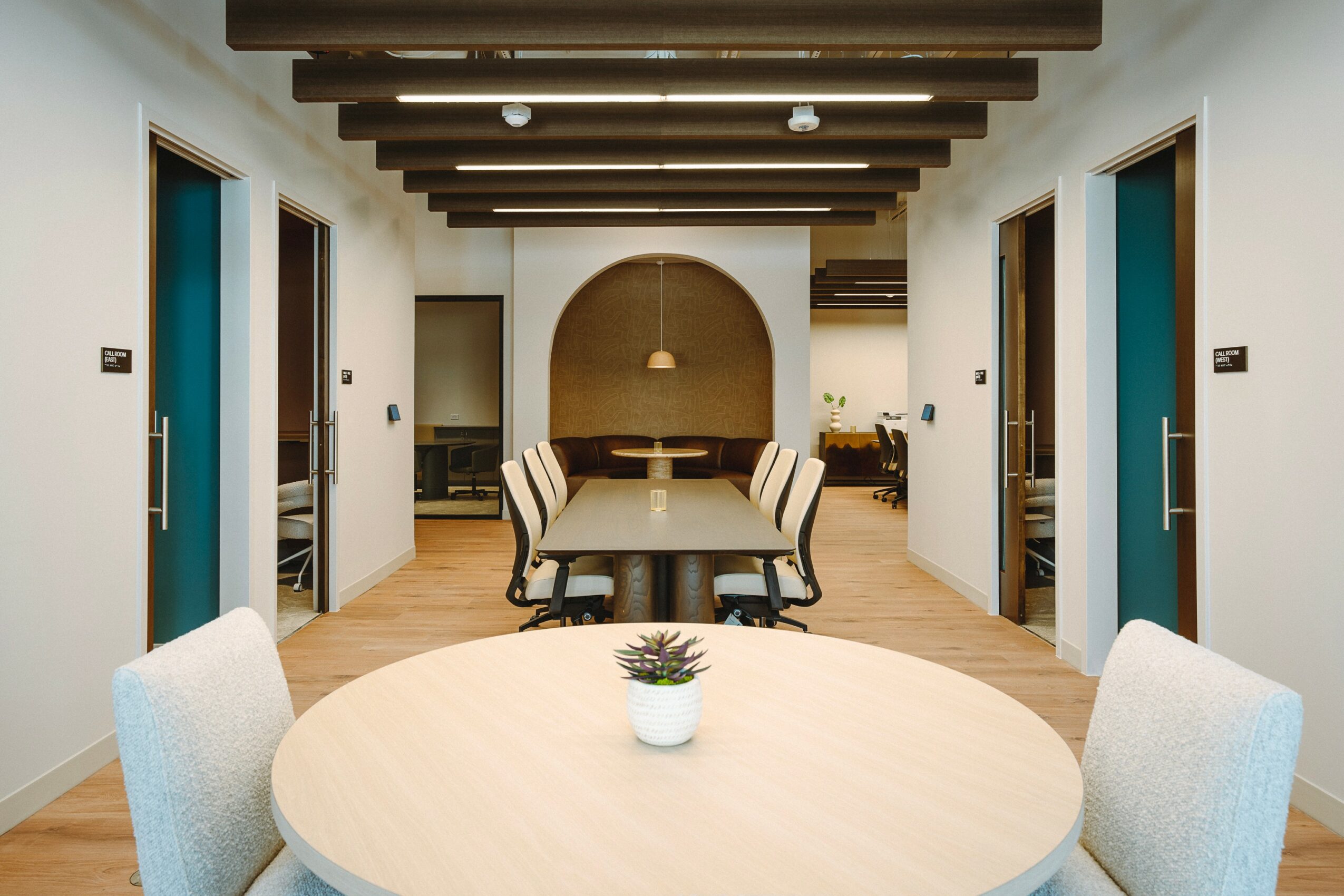 Modern conference room with a round table, chairs, and a hallway with doors on both sides, ceiling beams, and a decorative arch.