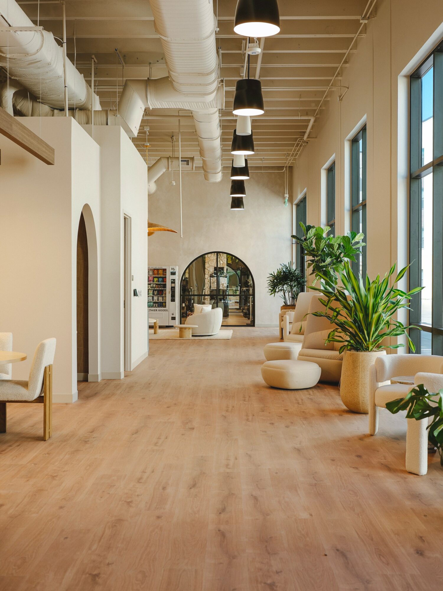 Long, open interior space with large windows, potted plants, seating, and high ceiling with exposed pipes and hanging lights.