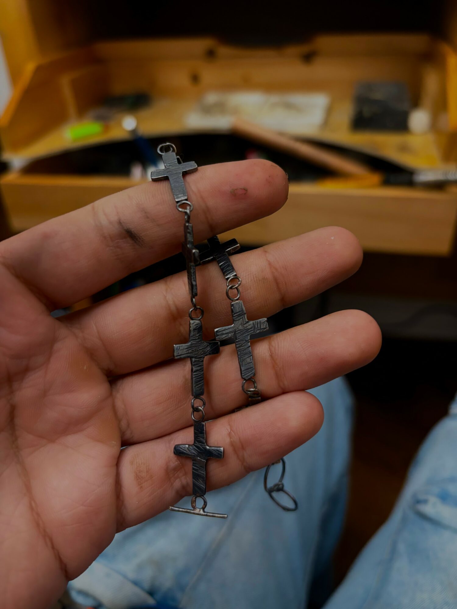 Hand holding a metal rosary with cross-shaped beads, background shows a yellow toolbox and a wooden surface.
