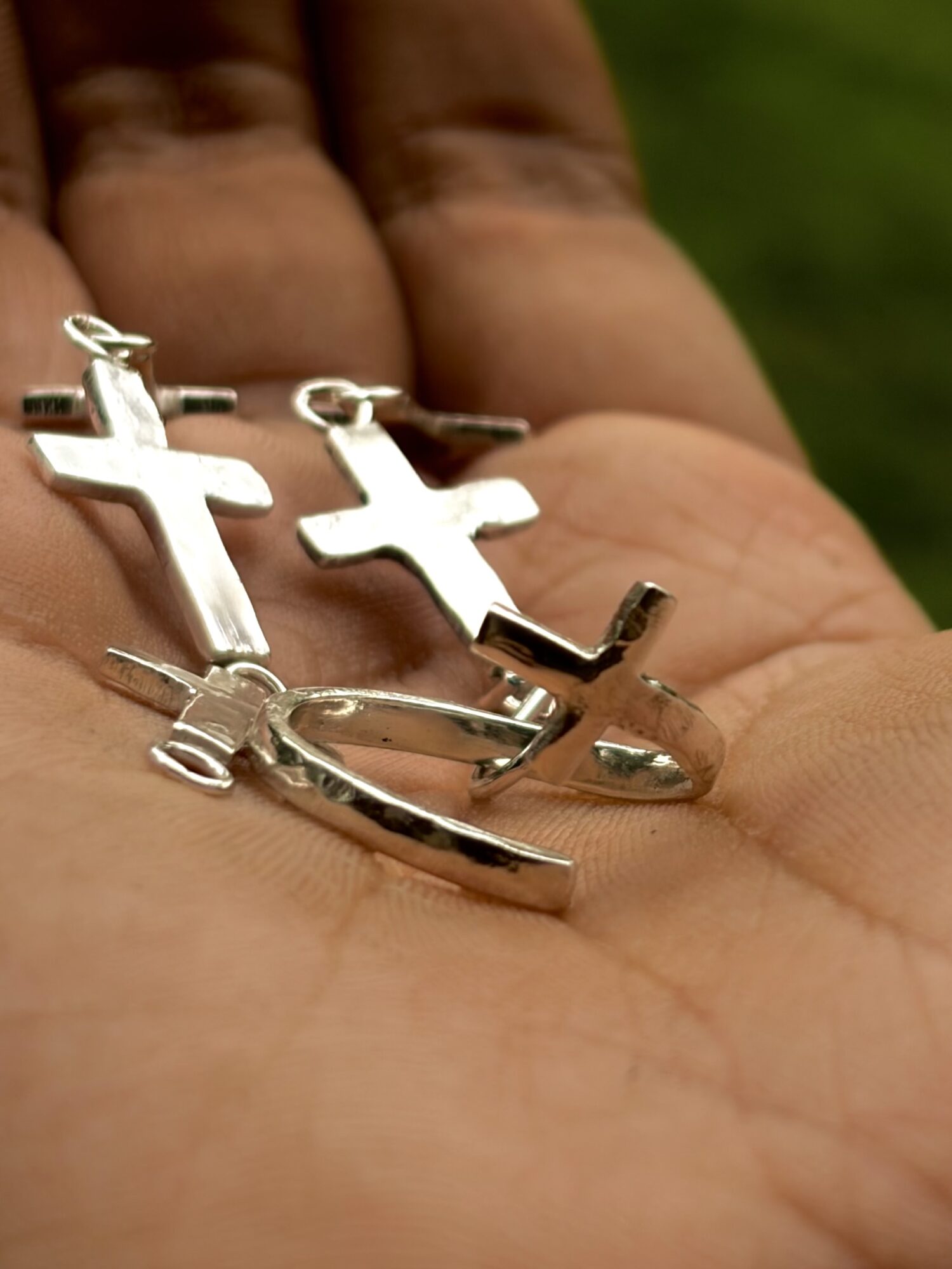 Hand holding three silver cross necklaces with different designs, against a blurred green background.