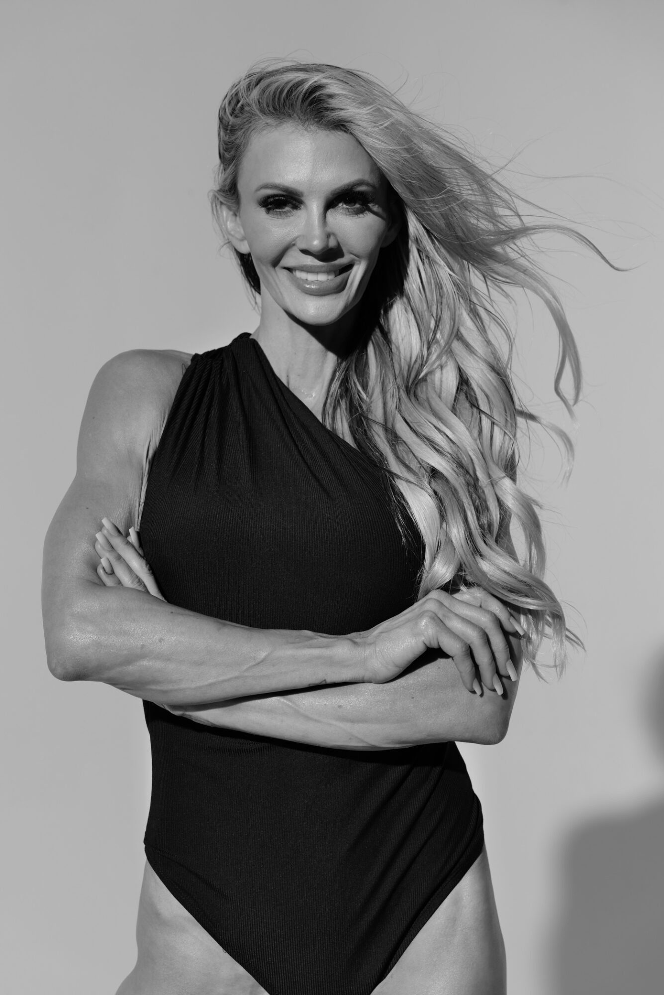 Woman with long, wavy hair smiling, arms crossed, wearing a sleeveless top, in black and white.