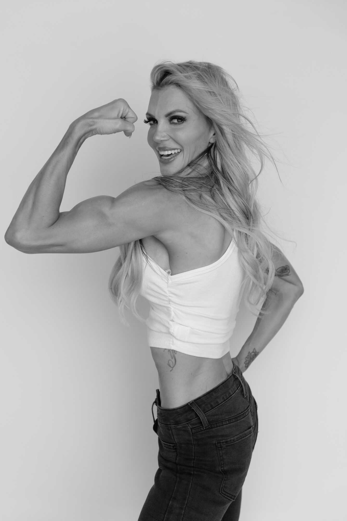 Woman with long hair flexing her arm, smiling, wearing a sleeveless top and jeans, standing sideways against a plain background.