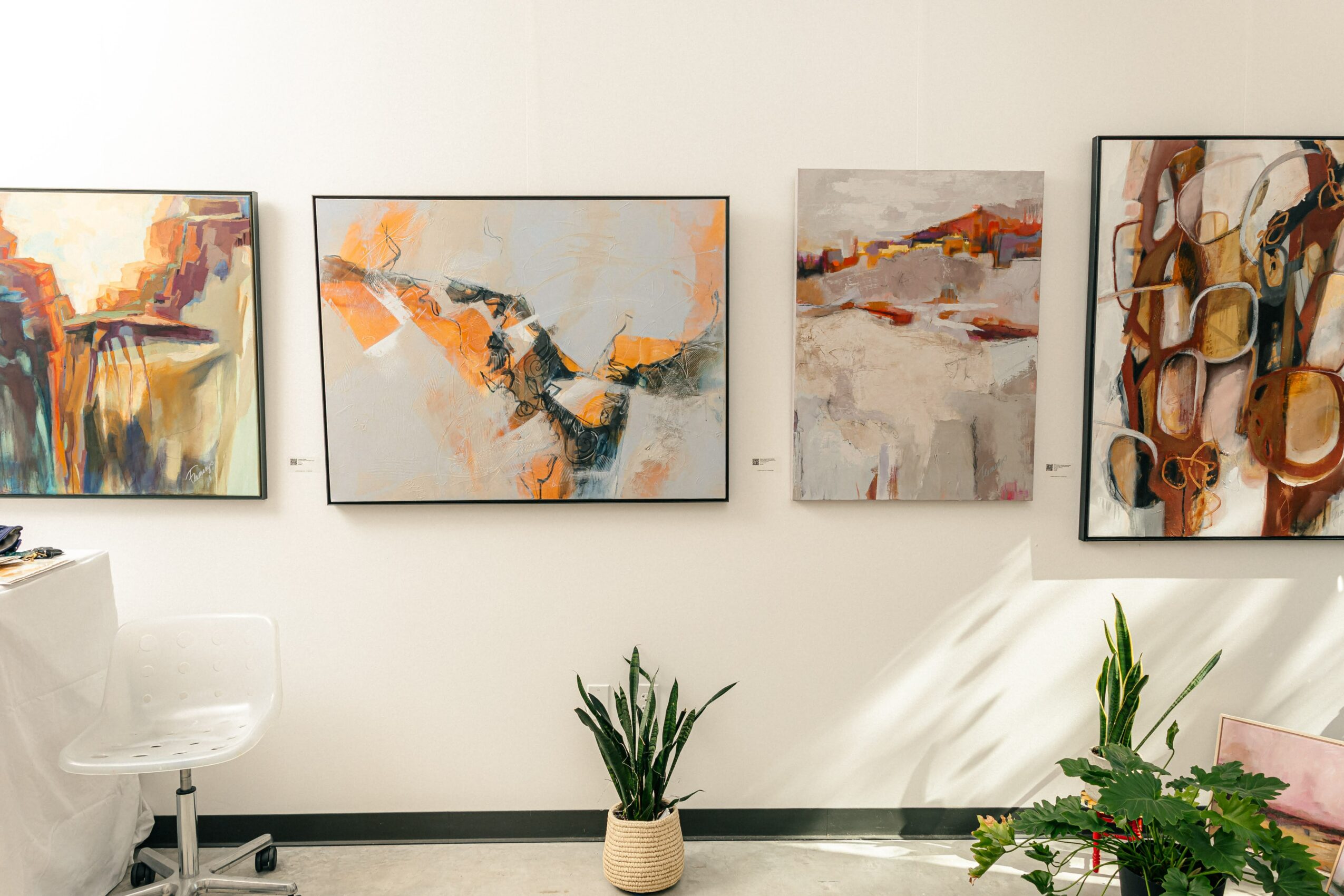 Four abstract paintings hang on a white wall, with two potted plants on the floor and sunlight casting shadows. Alt text 27 words.