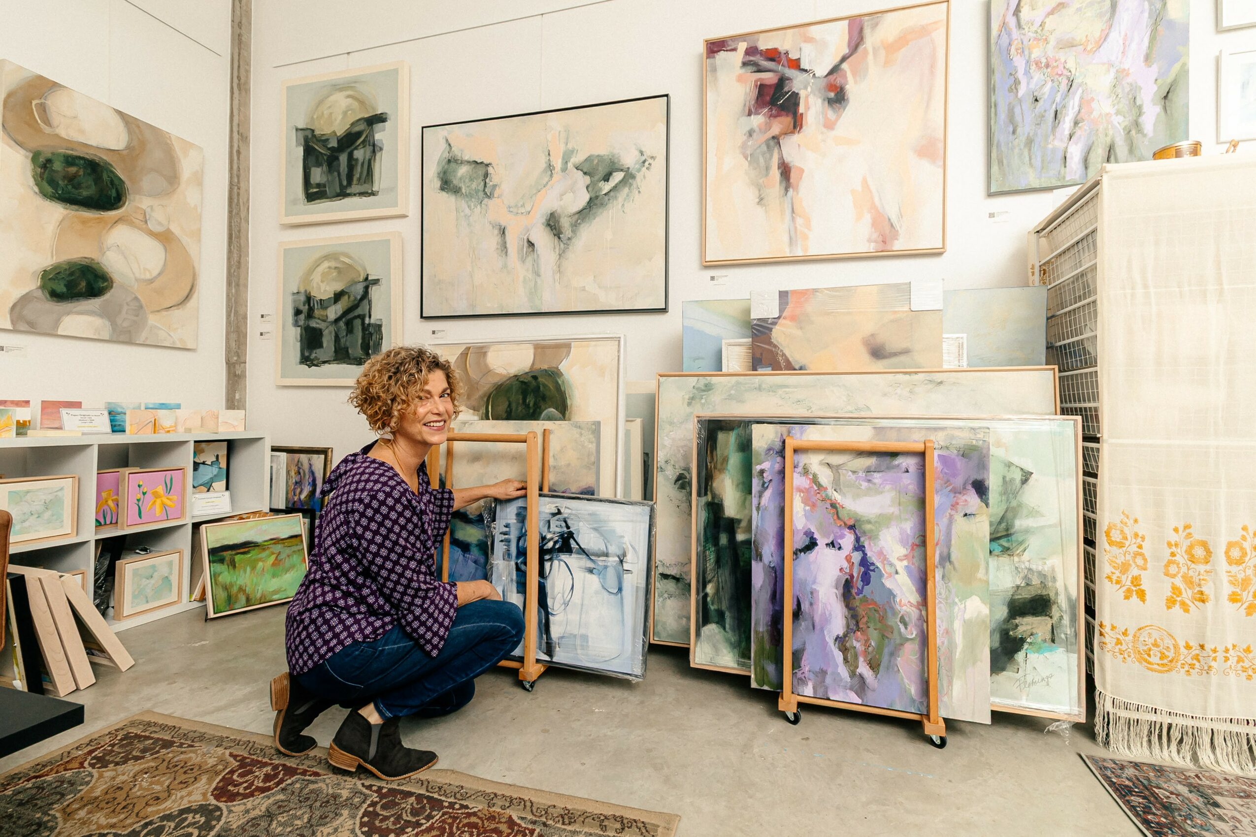 Woman with curly hair kneeling in art gallery surrounded by paintings and artwork on walls and shelves.