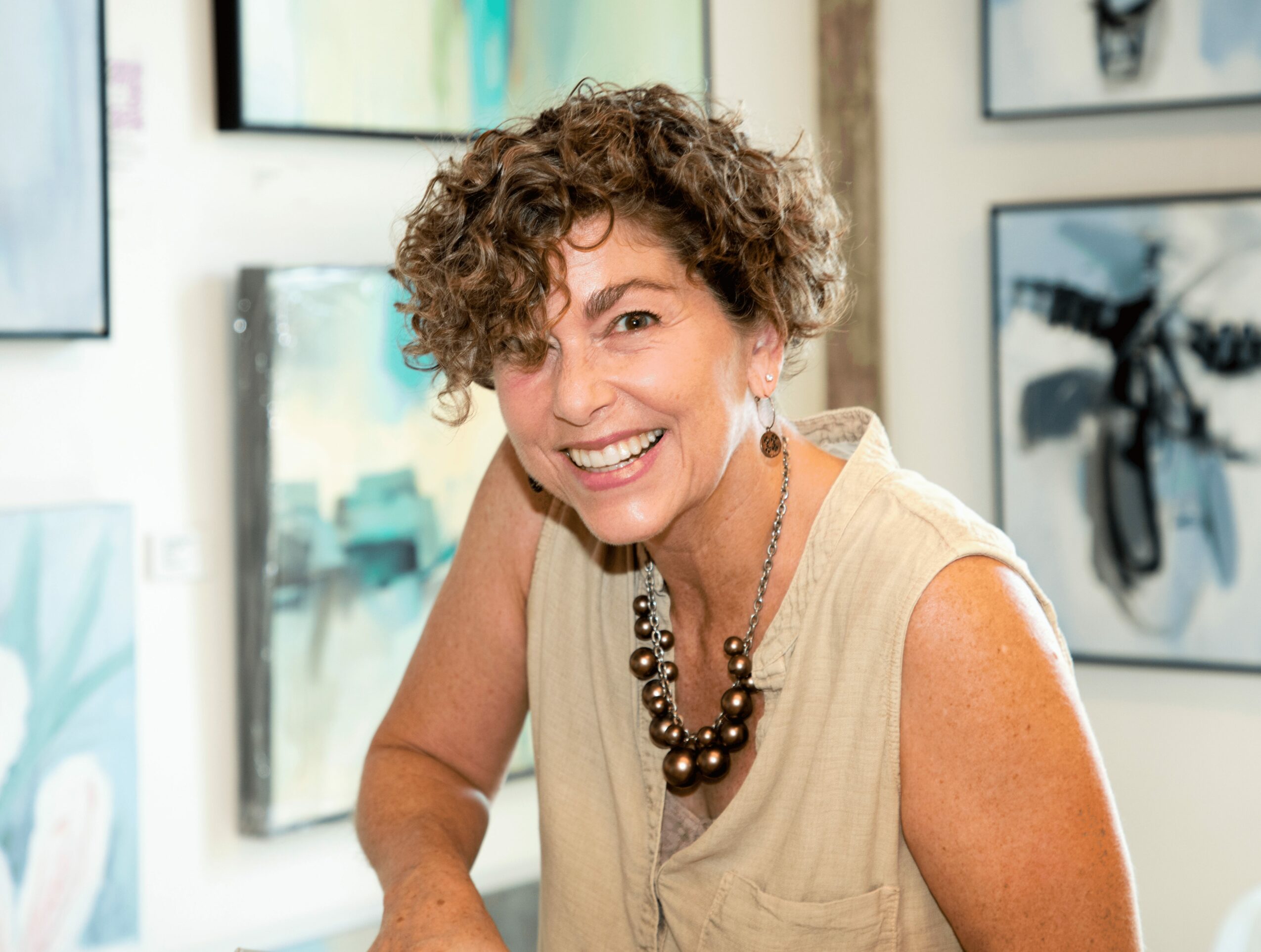 Woman with curly hair smiling, wearing a sleeveless beige top and a chunky necklace, in an art gallery.