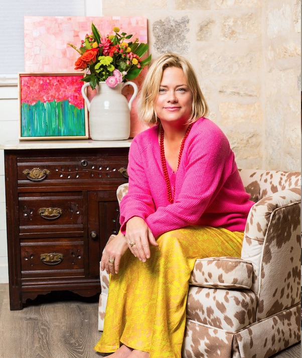 Woman with blonde hair in pink sweater and yellow pants sitting on patterned armchair, with colorful paintings and flowers behind her.