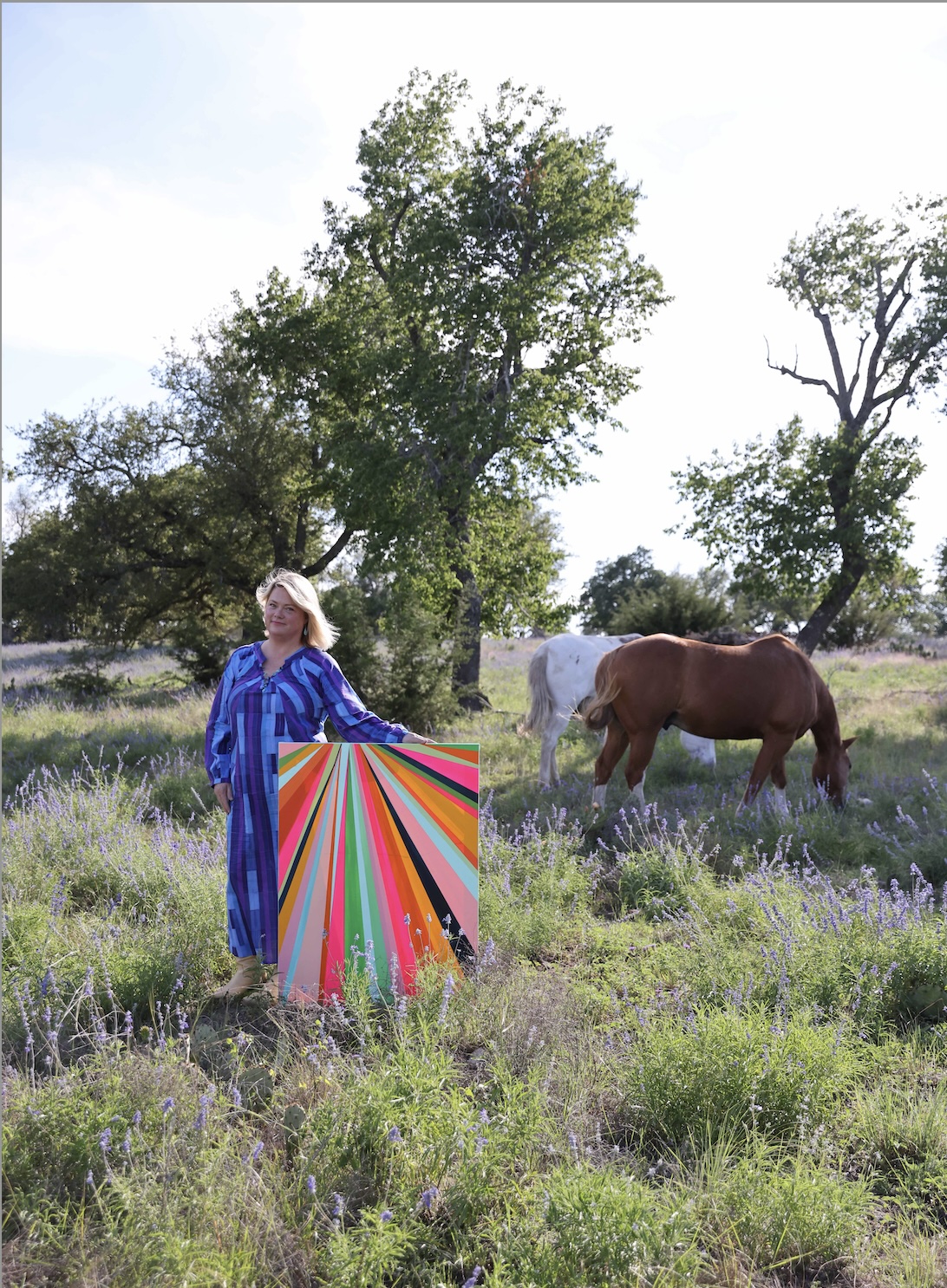 Woman in a blue dress holding a colorful umbrella in a grassy field with trees and a horse in the background.