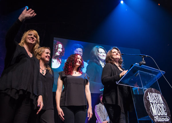 Four women stand on stage, one speaking at a clear podium with a logo, with a large screen behind them showing their images.