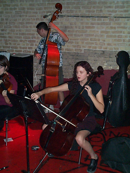 Young girl playing cello on stage with other musicians, brick wall background, red carpet, music stand, and instrument cases nearby.